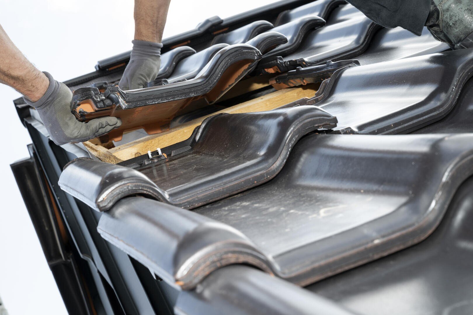 Nahaufnahme von zwei Arbeitern, die mit Handschuhen dunkle Keramikdachziegel auf einem Dach verlegen, wobei unter den Ziegeln Holzbalken sichtbar sind.