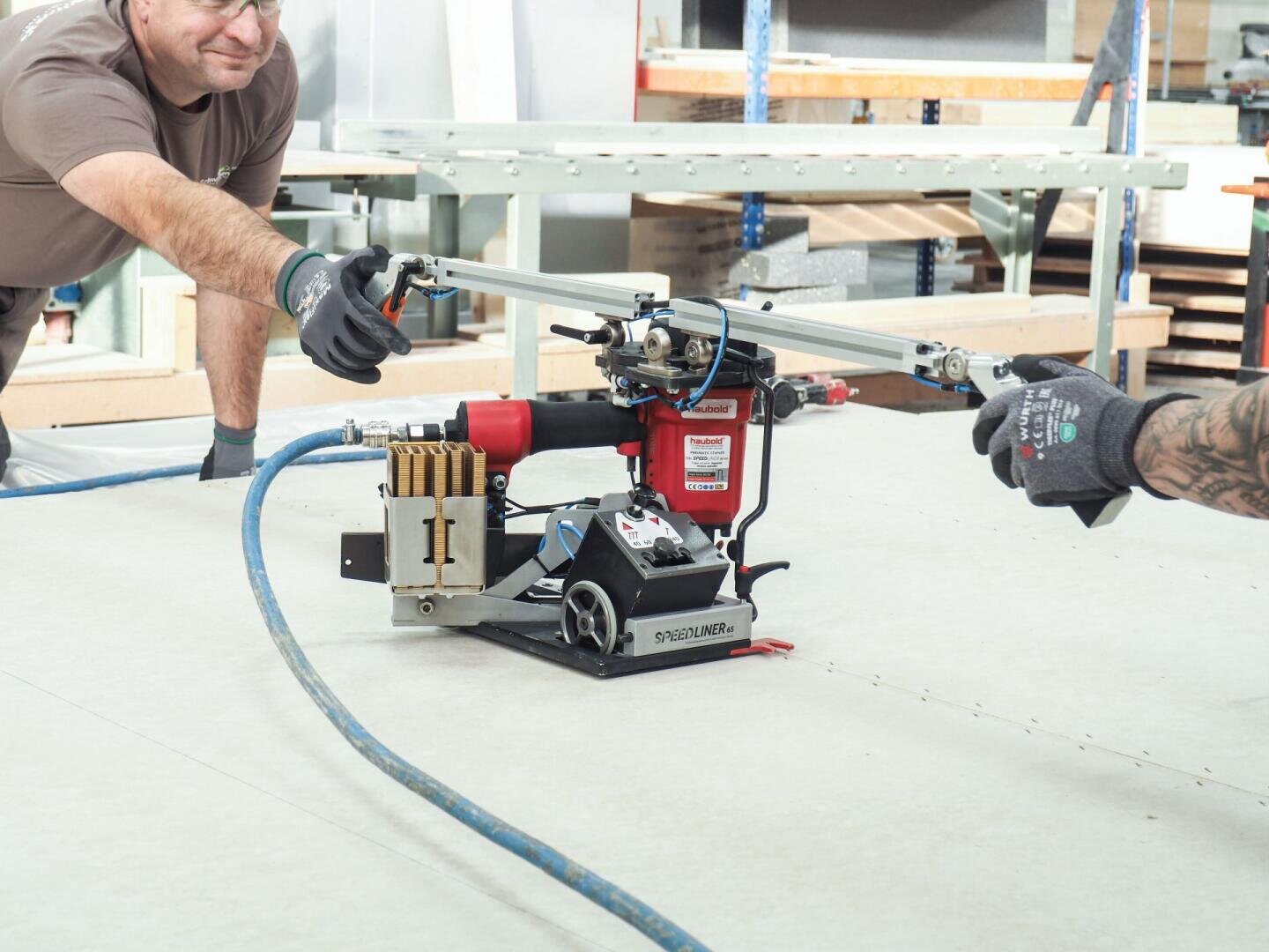 Zwei Personen bedienen eine spezielle Bodenheftmaschine mit einem verlängerten Griff auf einer hellen Fläche in einer Werkstatt, beide tragen Handschuhe und legere Arbeitskleidung. Im Hintergrund sind Regale und Vorräte zu sehen.