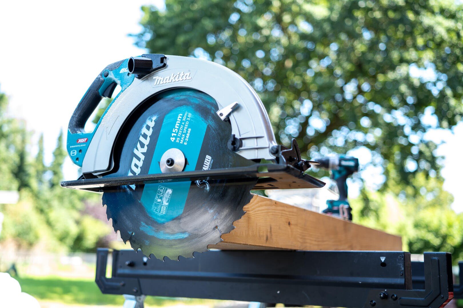 Eine blaue Makita-Kreissäge liegt auf einem Holzbrett im Freien, mit Bäumen und Grünzeug im Hintergrund an einem sonnigen Tag.