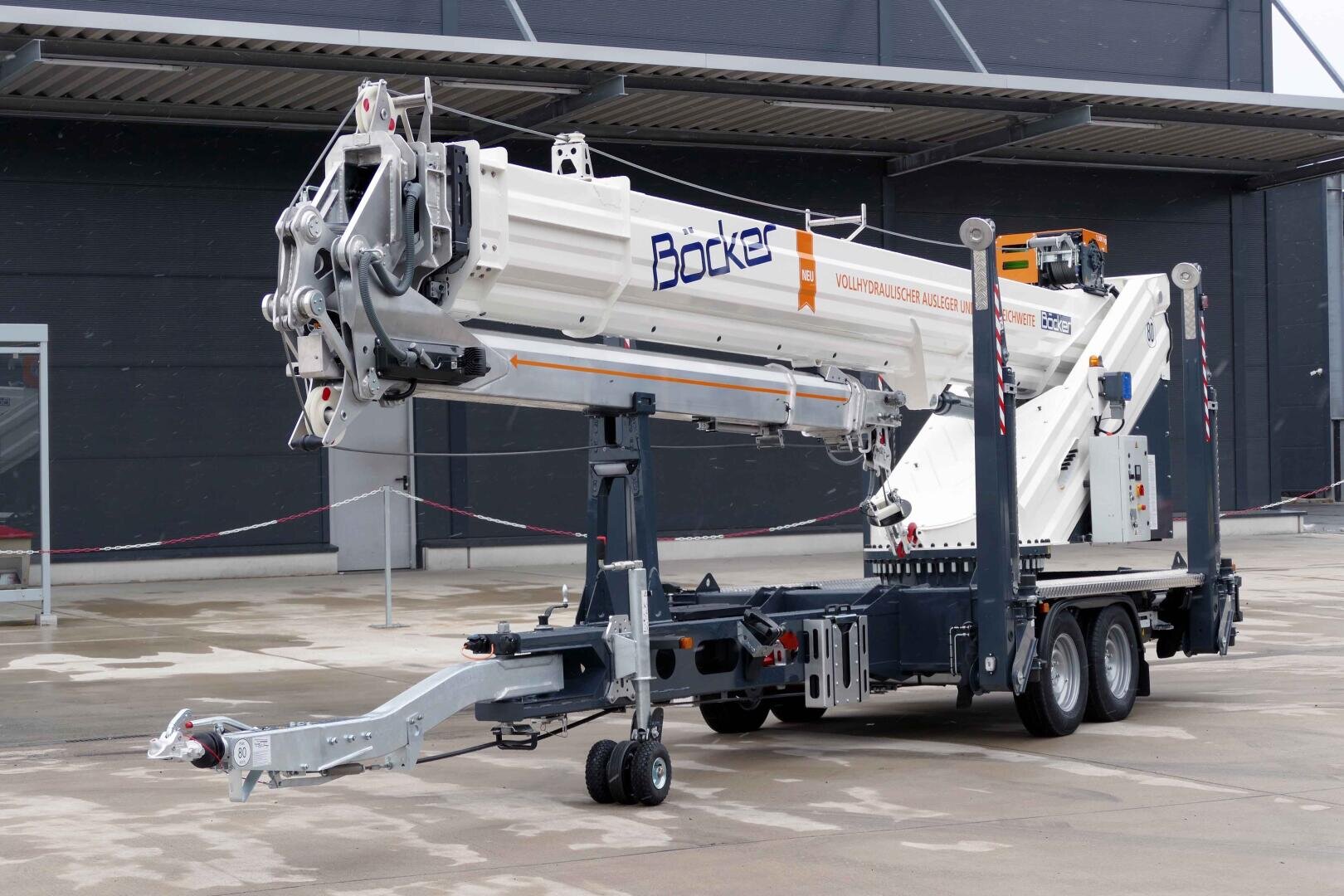 A Böcker mobile construction crane is parked on a paved area in front of a building. The crane is mounted on a trailer with extendable supports and appears to be ready for transportation.