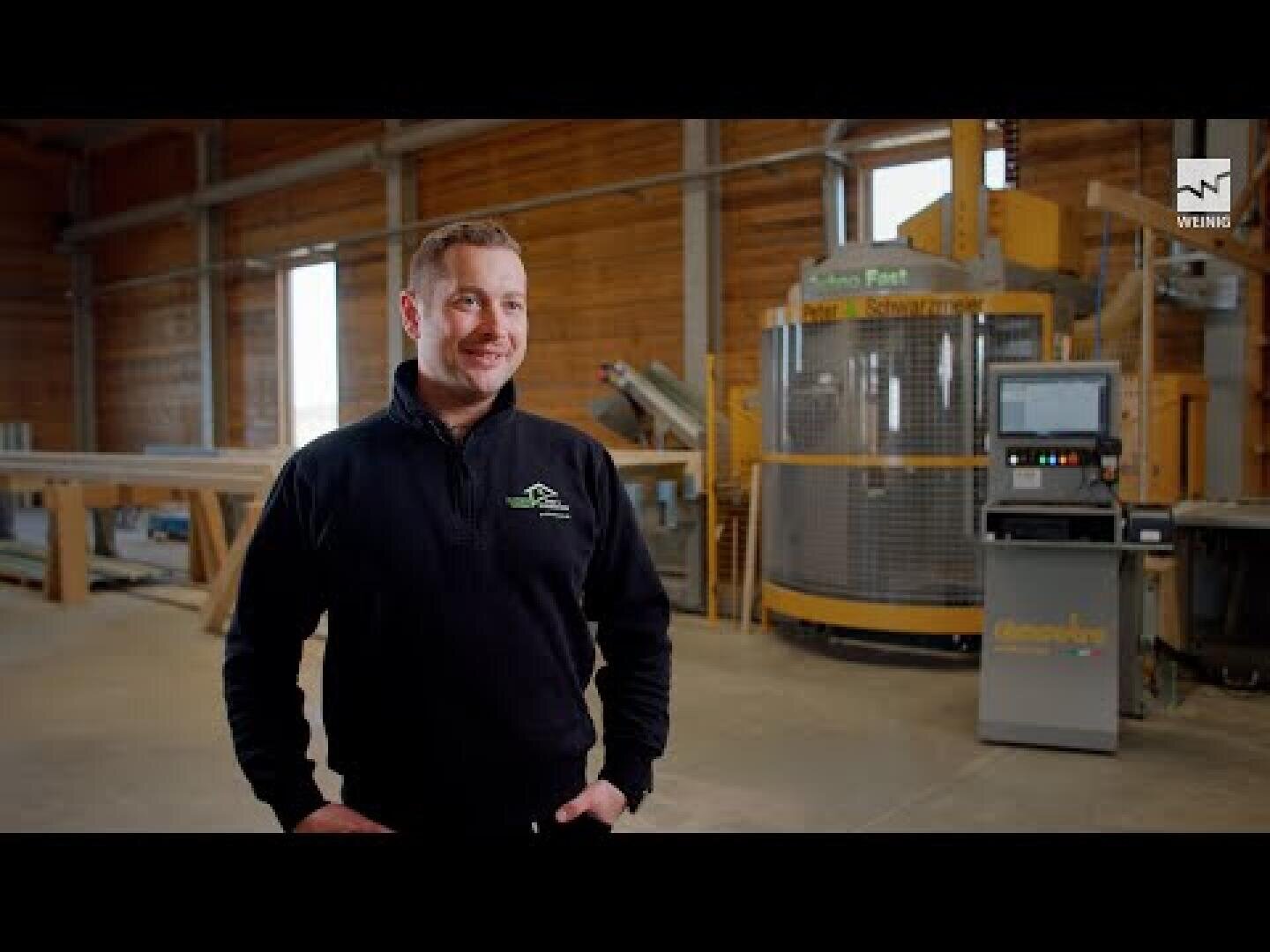 Ein Mann in einem dunklen Pullover steht lächelnd in einer geräumigen, gut beleuchteten Industriehalle mit Holzwänden und großen Maschinen im Hintergrund.