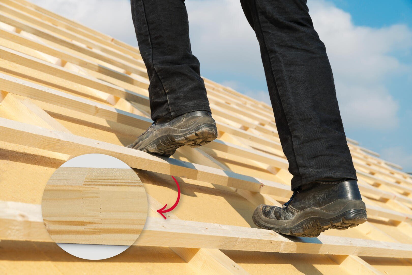 A person in black pants and work boots stands on a wooden roof truss. An inset circle with a red arrow highlights a close-up of the wood grain and structure. The sky is blue with some clouds.