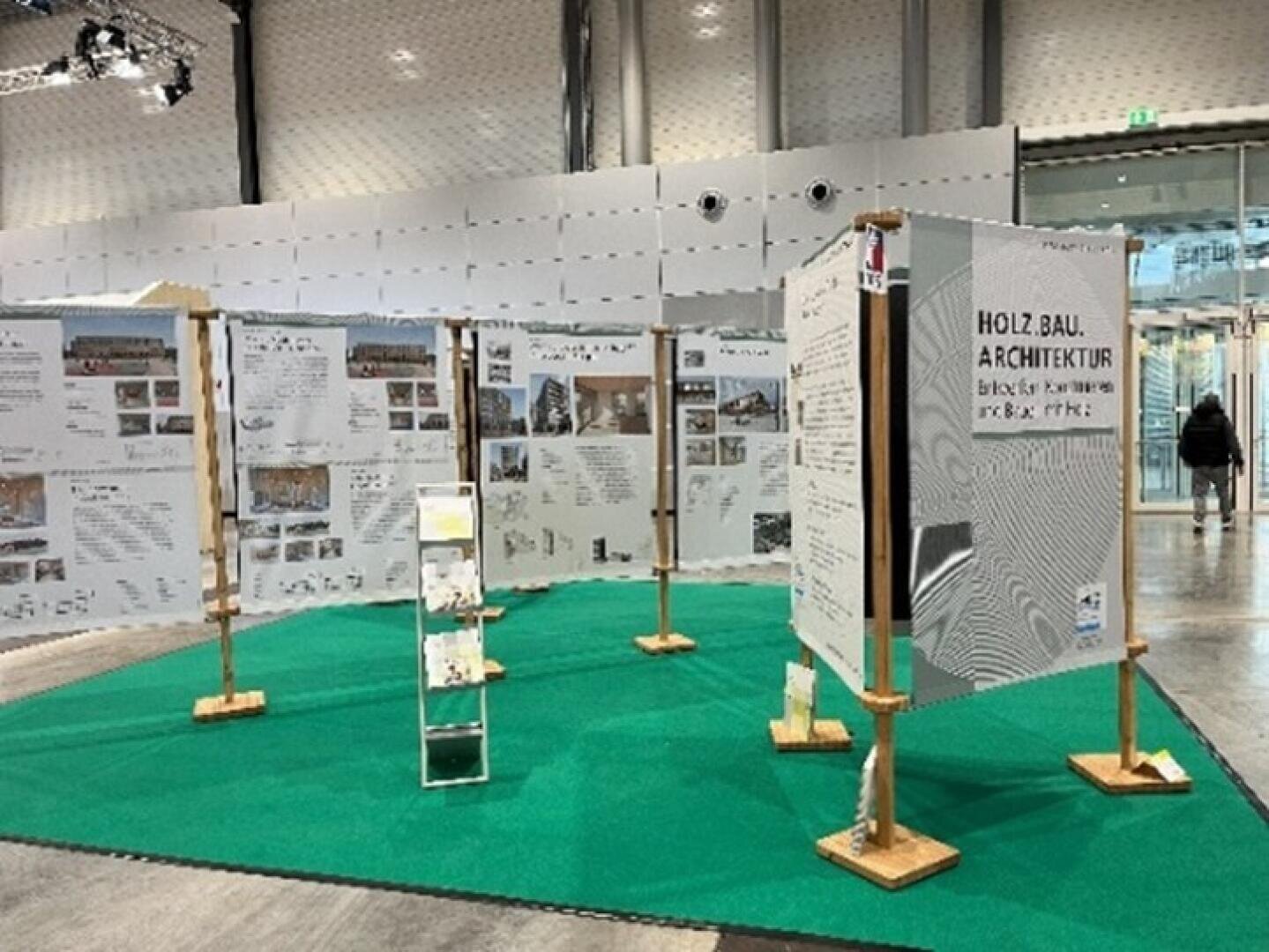 Ausstellung mit großen, bedruckten Tafeln auf Holzständern mit Informationen und Bildern über Holzbau und Architektur, auf einem grünen Teppich in einer geräumigen Halle.