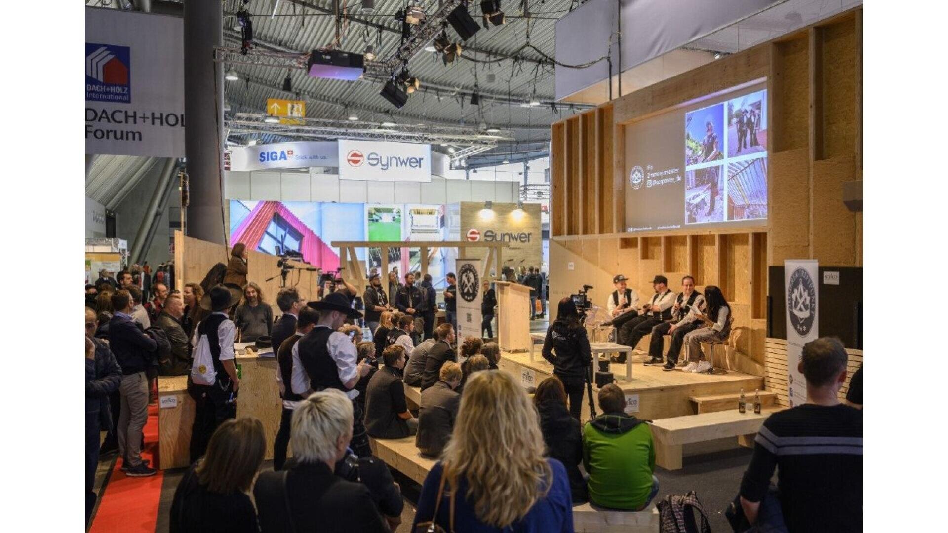 A crowd attends a presentation at a trade show inside a large hall. People are seated and standing, facing speakers on a stage with wood displays and large screens showing images and logos. Exhibition booths are visible in the background.