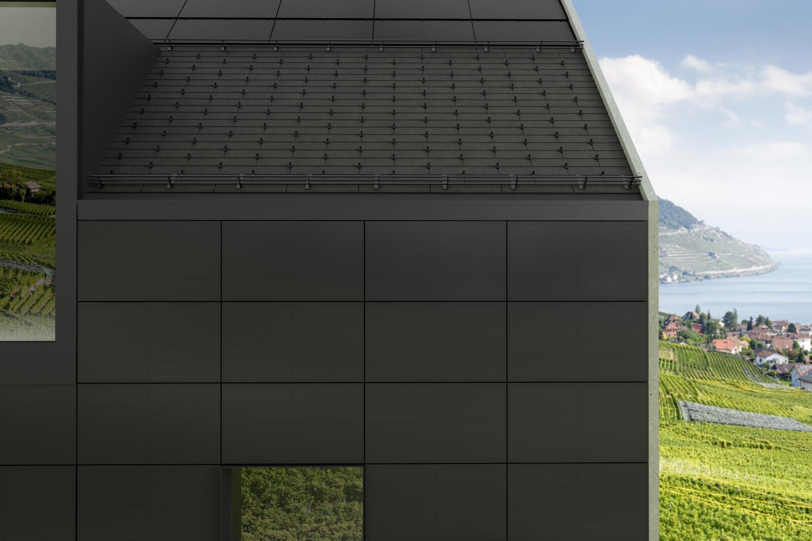 Modernes schwarzes Gebäude mit Solarzellen auf dem Dach, großen Fliesen an der Außenseite und einem Fenster, in dem sich grüne Weinberge spiegeln. Im Hintergrund sind sanfte Hügel, Weinberge, Häuser und ein blauer See unter einem teilweise bewölkten Himmel zu sehen.