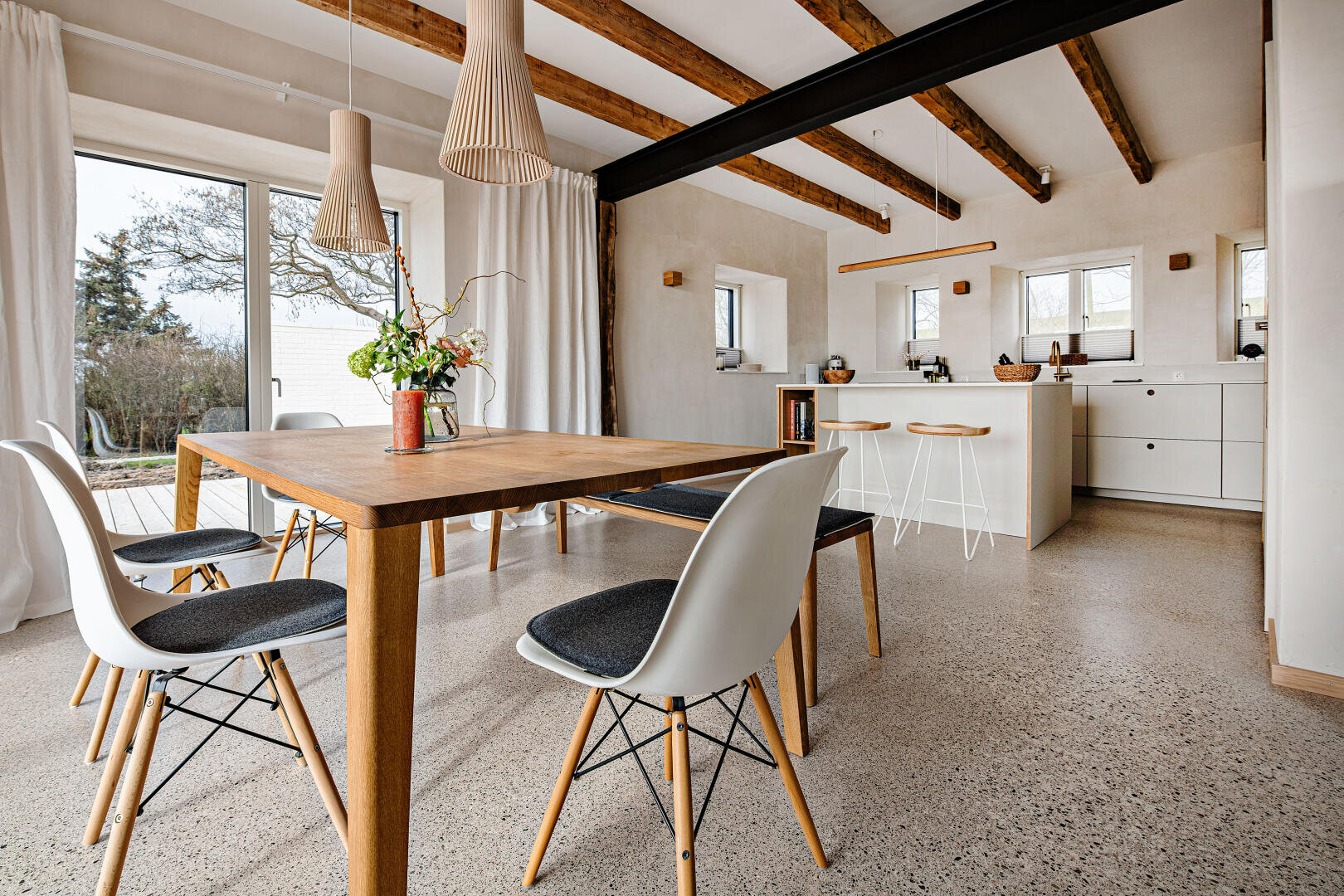 Ein moderner Ess- und Küchenbereich mit Holzbalken, großen Fenstern, einem Holztisch mit weißen Stühlen, Pendelleuchten, minimalistischen weißen Schränken und einem Blick auf die Bäume draußen.
