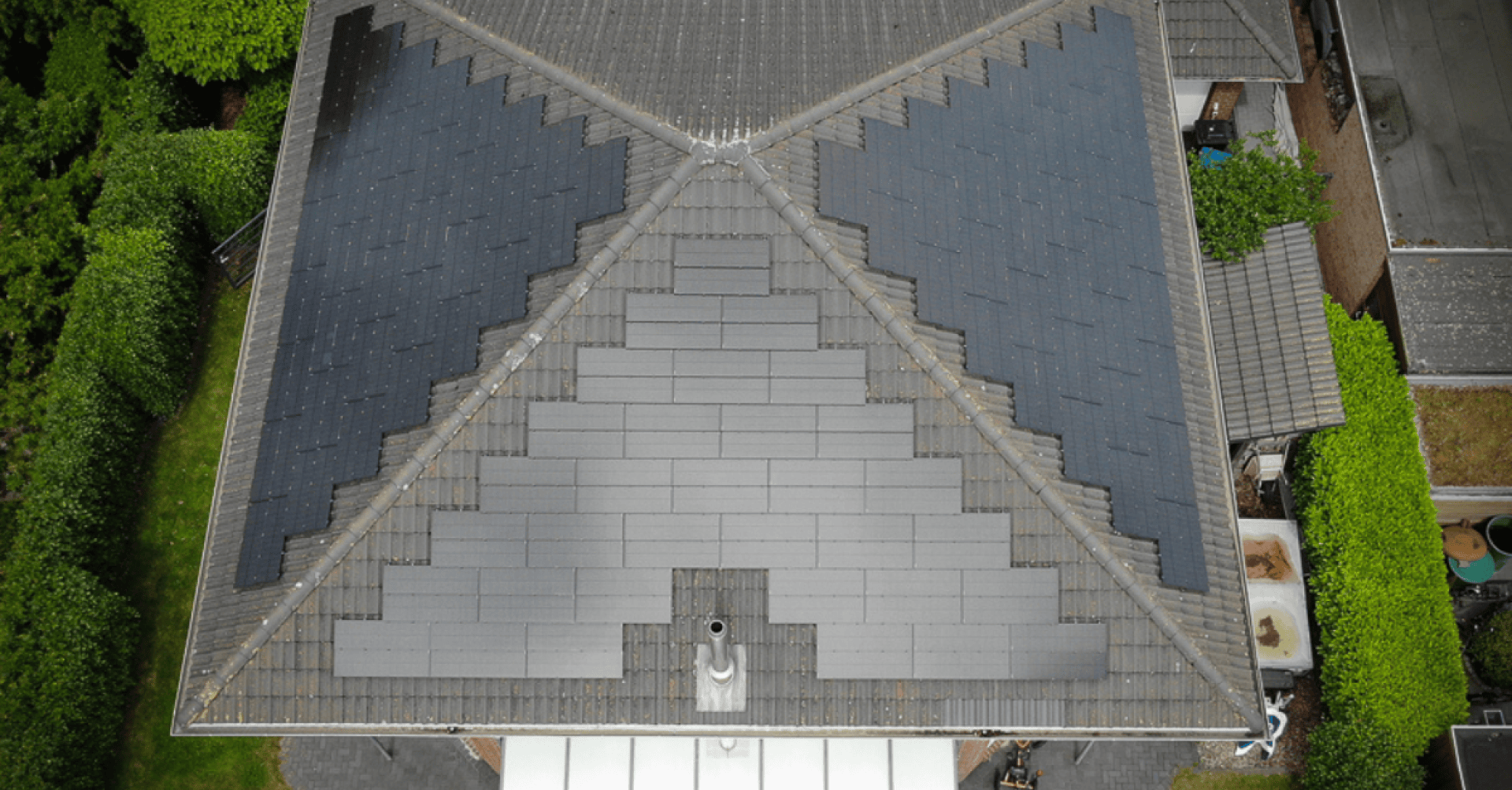 Luftaufnahme eines Hausdachs mit einer Reihe von schwarzen Solarmodulen, die in einem symmetrischen, geometrischen Muster angeordnet sind, umgeben von Bäumen und benachbarten Gebäuden.