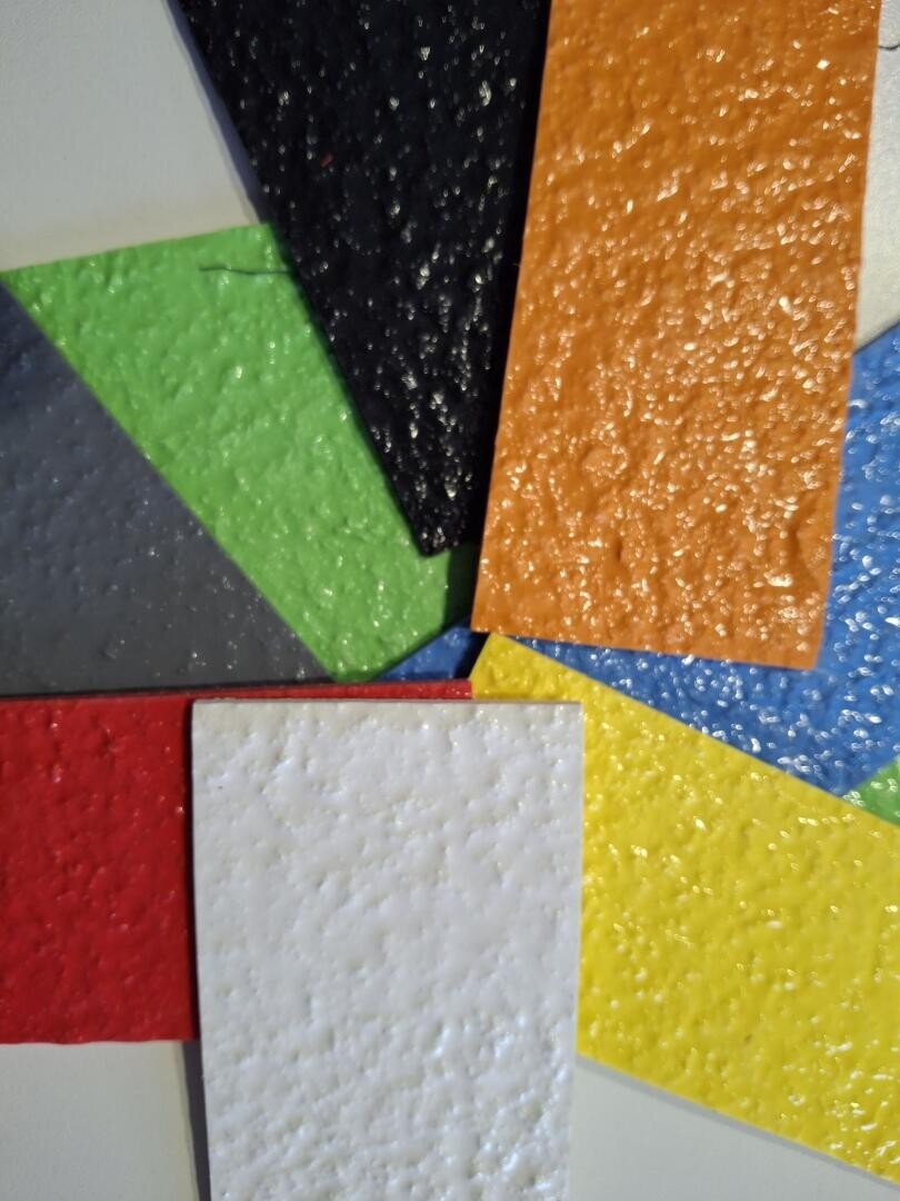 Neun strukturierte rechteckige Kunststoffplatten in verschiedenen Farben (schwarz, orange, blau, gelb, weiß, rot, grün, hellgrau und dunkelgrau) sind fächerförmig auf einer ebenen Fläche angeordnet.