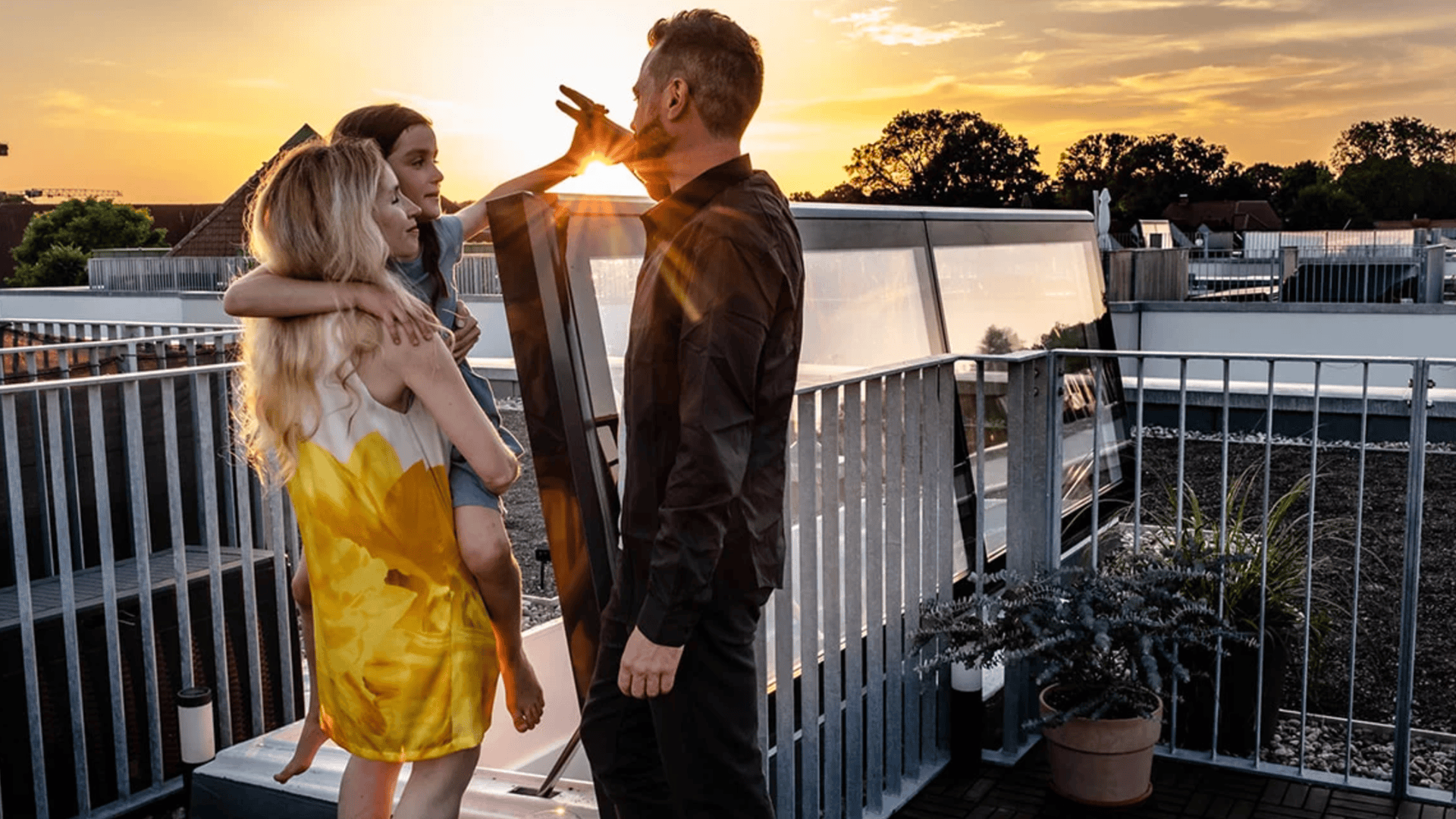 Ein Mann, eine Frau und ein junges Mädchen stehen bei Sonnenuntergang gemeinsam auf einer Dachterrasse. Die Frau hält das Mädchen im Arm, der Mann streckt die Hand nach ihr aus. Im Hintergrund sind moderne Gebäude und Bäume zu sehen.