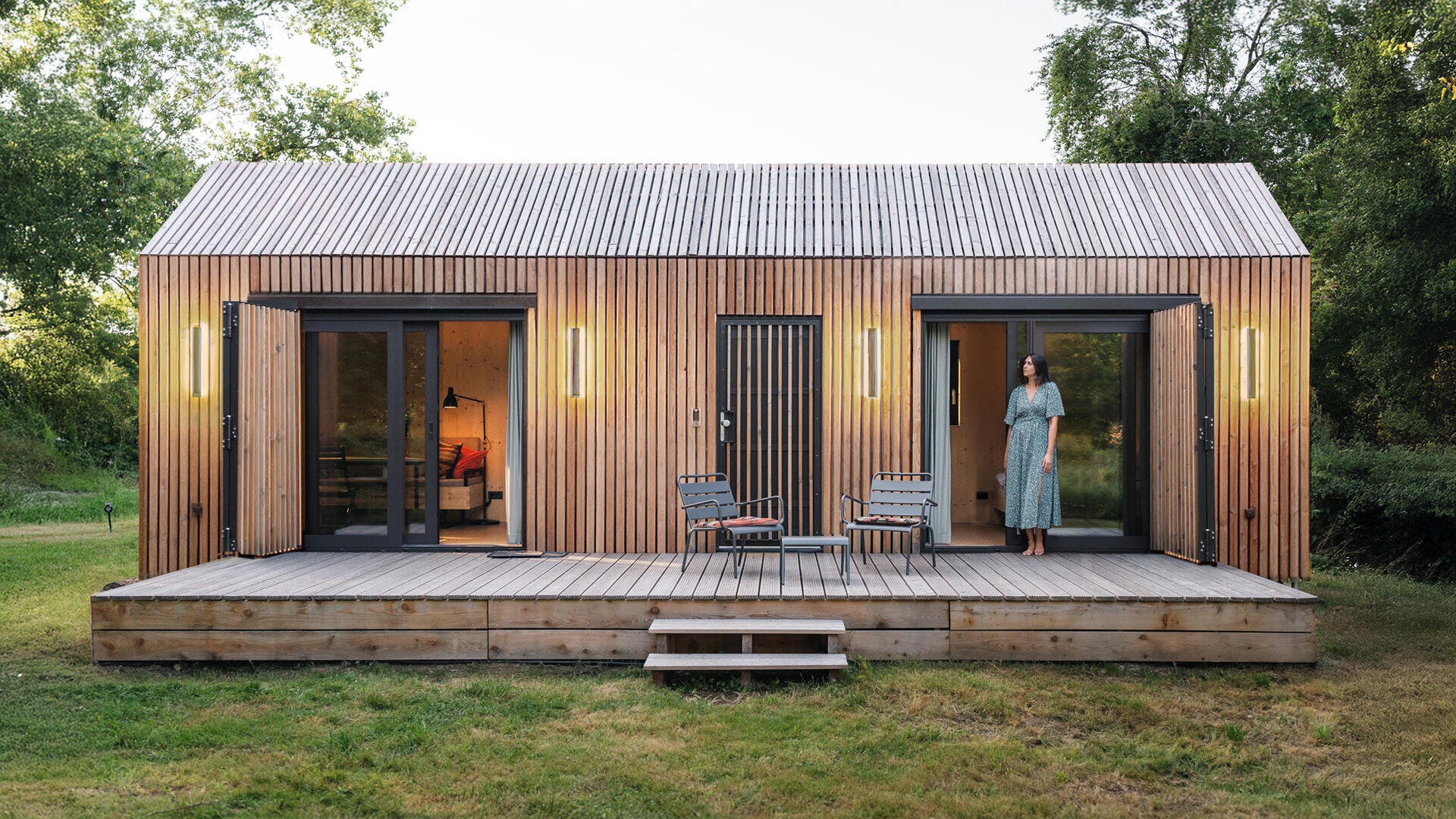 Ein modernes kleines Haus mit vertikaler Holzverkleidung, großen Glastüren und einer kleinen Veranda mit zwei Stühlen und einem Tisch. Eine Frau in einem langen Kleid steht auf der Veranda. Das Haus ist von grünen Bäumen und Gras umgeben.