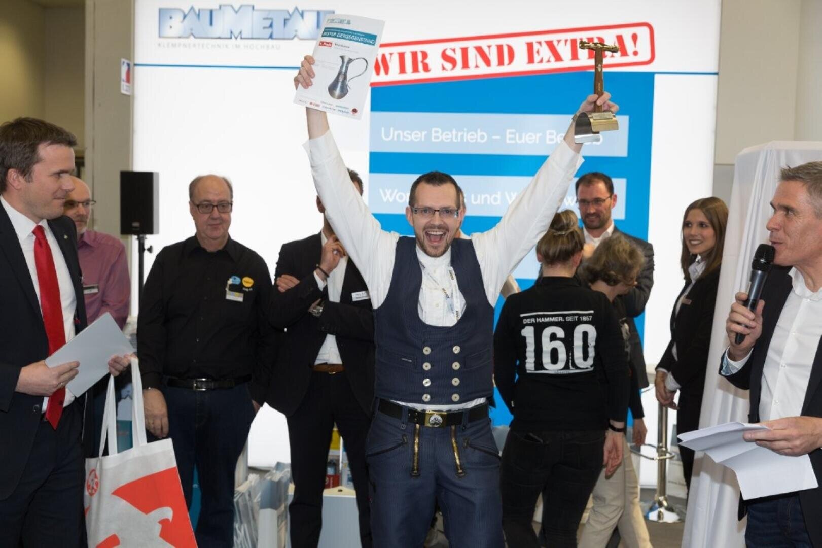 A man in traditional dress celebrates on stage, holding a certificate and a trophy above his head, surrounded by smiling people at an awards ceremony. A large sign and several applauding onlookers can be seen in the background.