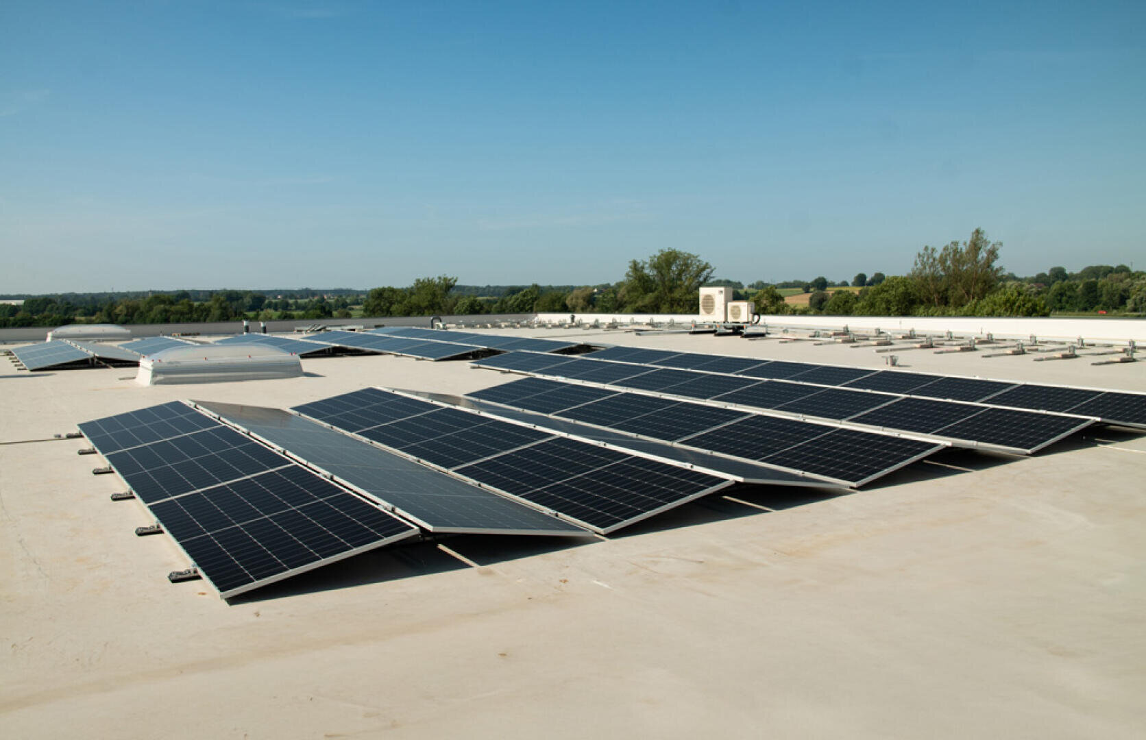 Auf einem Flachdach installierte Solarmodule unter blauem Himmel, mit grünen Bäumen und Feldern im Hintergrund.