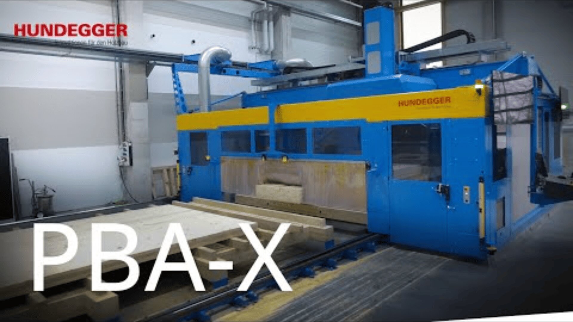 Eine große blau-gelbe Hundegger PBA-X Industrie-Holzbearbeitungsmaschine bearbeitet Holzbalken in einer modernen Fabrik. Die Maschine ist von Metallschienen und Fenstern umgeben, in der Ecke prangt das Hundegger-Logo.