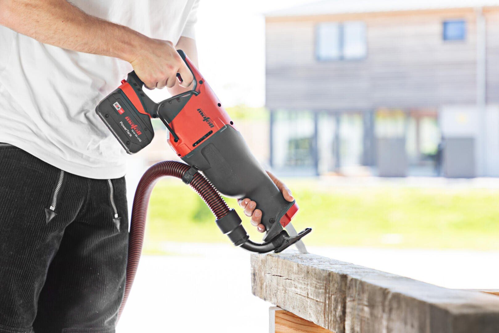 Eine Person in einem weißen Hemd benutzt eine rot-schwarze elektrische Säbelsäge mit einem Absaugschlauch, um im Freien ein Holzbrett zu durchtrennen, mit einem unscharfen Gebäude im Hintergrund.