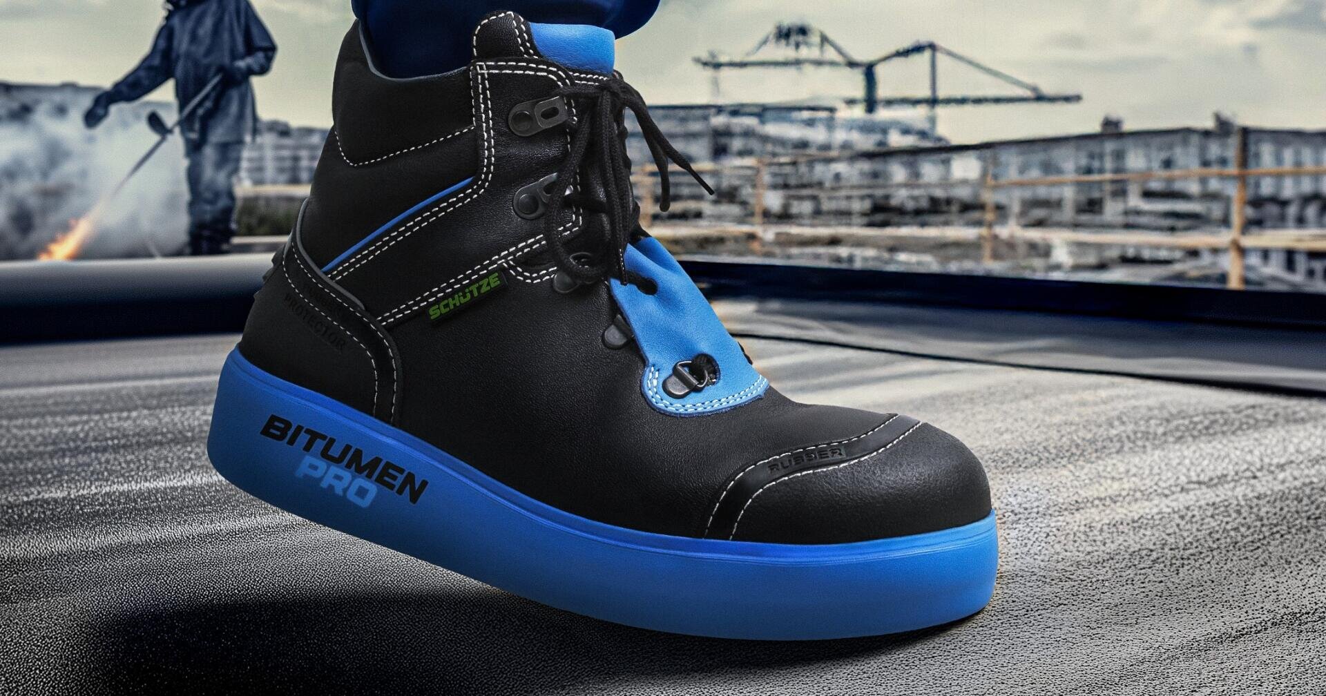 Eine Nahaufnahme eines schwarz-blauen Sicherheitsstiefels mit der Aufschrift BITUMEN PRO und einer industriellen Baustelle im Hintergrund.
