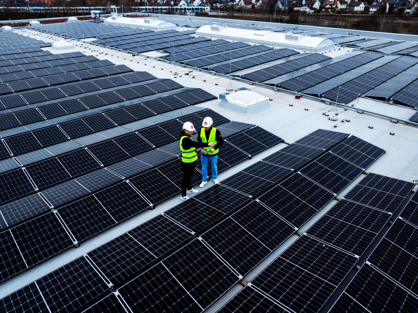Zwei Personen in Sicherheitswesten und Helmen stehen zwischen Reihen von Solarmodulen auf einem Dach und besprechen oder überprüfen die Installation. Die Paneele bedecken den größten Teil des Flachdachs, im Hintergrund ist eine Stadt zu sehen.