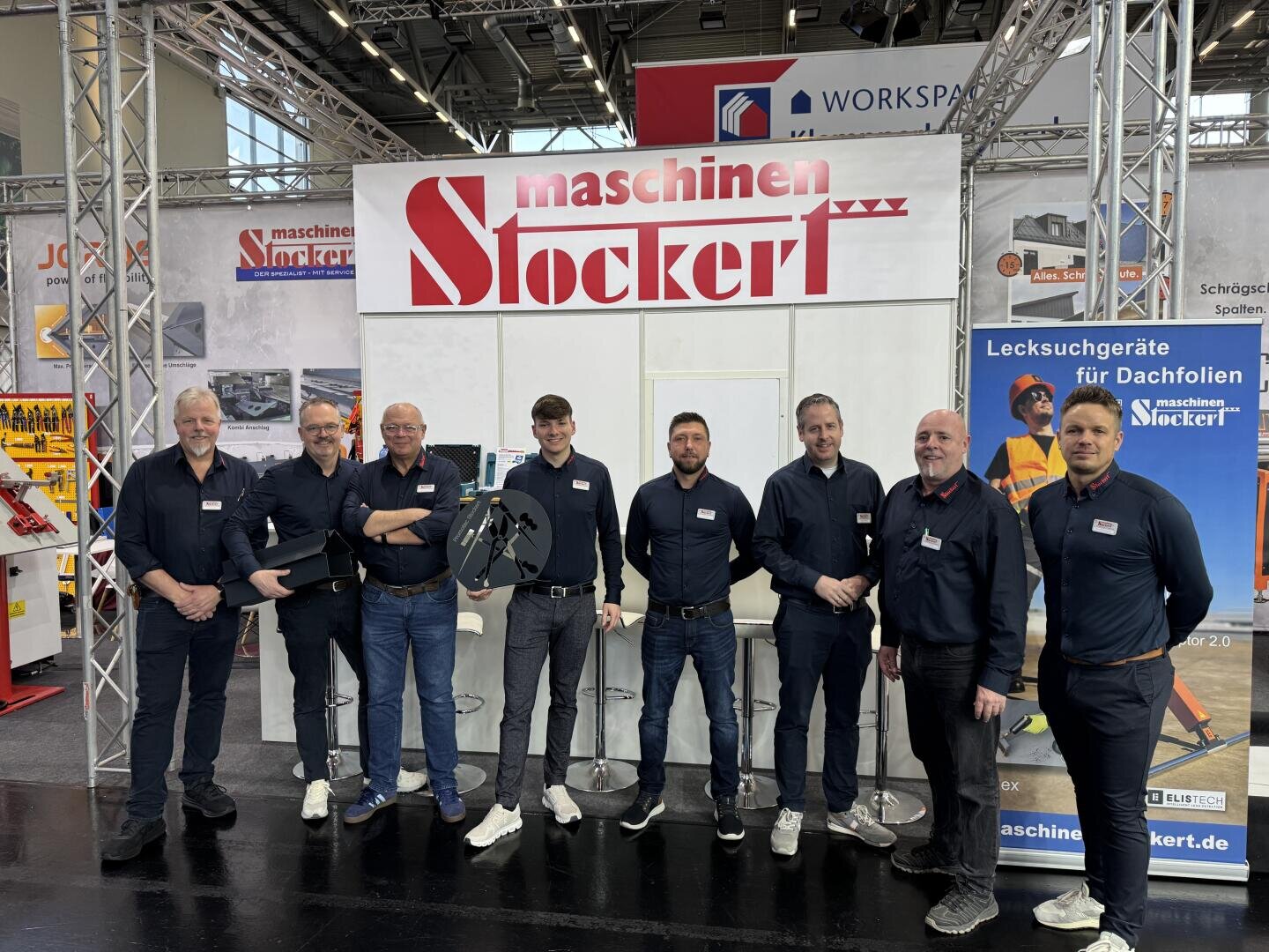 Acht Männer mit dunklen Hemden und Namensschildern stehen lächelnd vor einem Stand von Maschinen Stockert auf einer Messe, während im Hintergrund Maschinen und Werbebanner zu sehen sind.