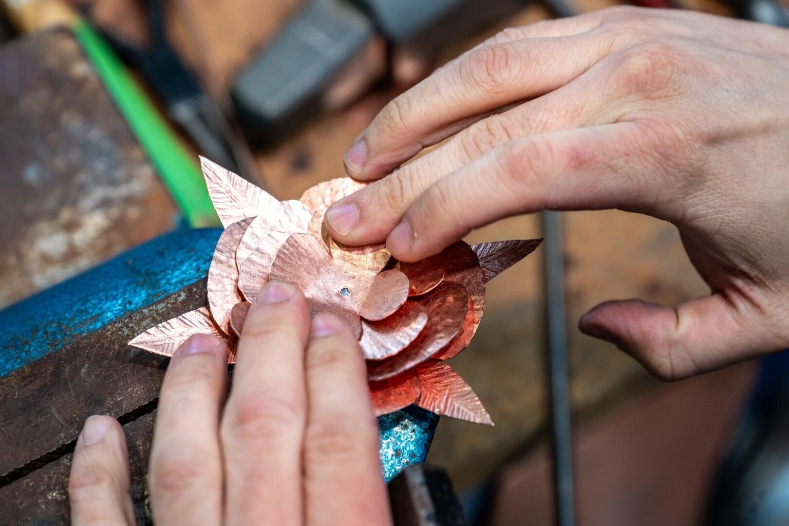 Die Hände einer Person fügen in einer Werkstatt Schichten von strukturierten Kupferblättern zusammen und formen ein blumenartiges Metallstück auf einem blauen Schraubstock, der von Werkzeugen umgeben ist.
