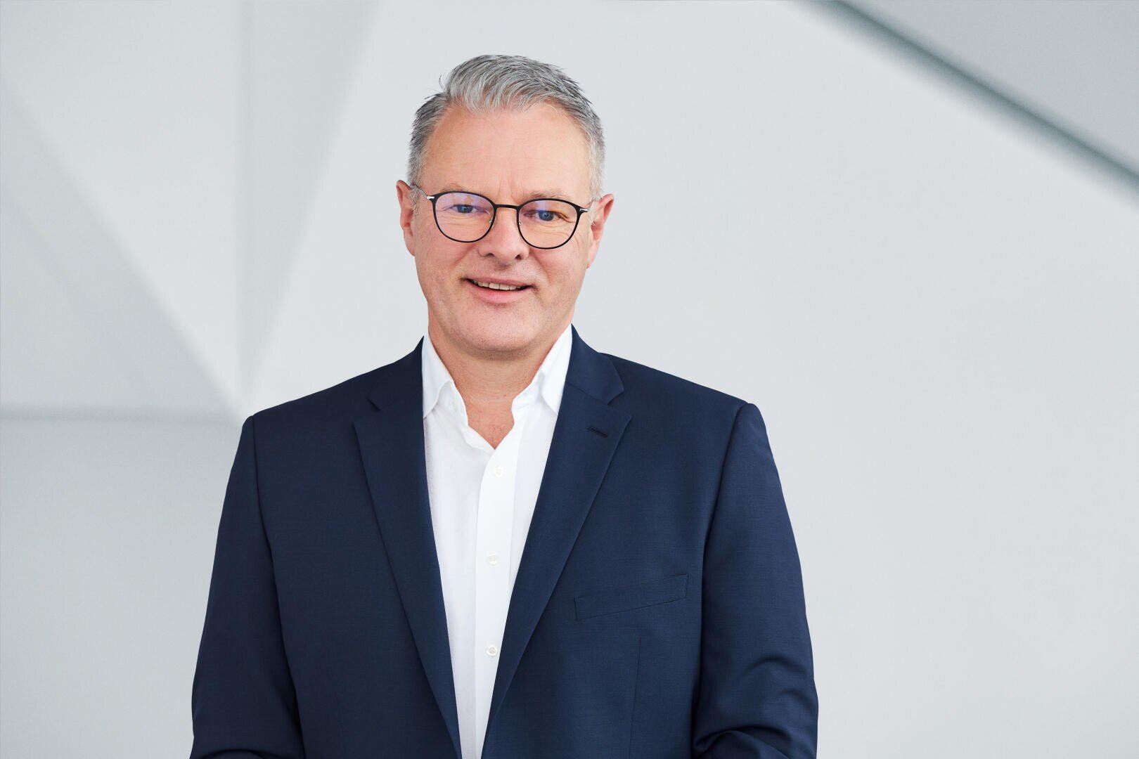 Ein Mann mittleren Alters mit kurzen grauen Haaren und Brille, der einen marineblauen Anzug und ein weißes Hemd trägt, steht vor einem hellgrauen, geometrischen Hintergrund und lächelt leicht.