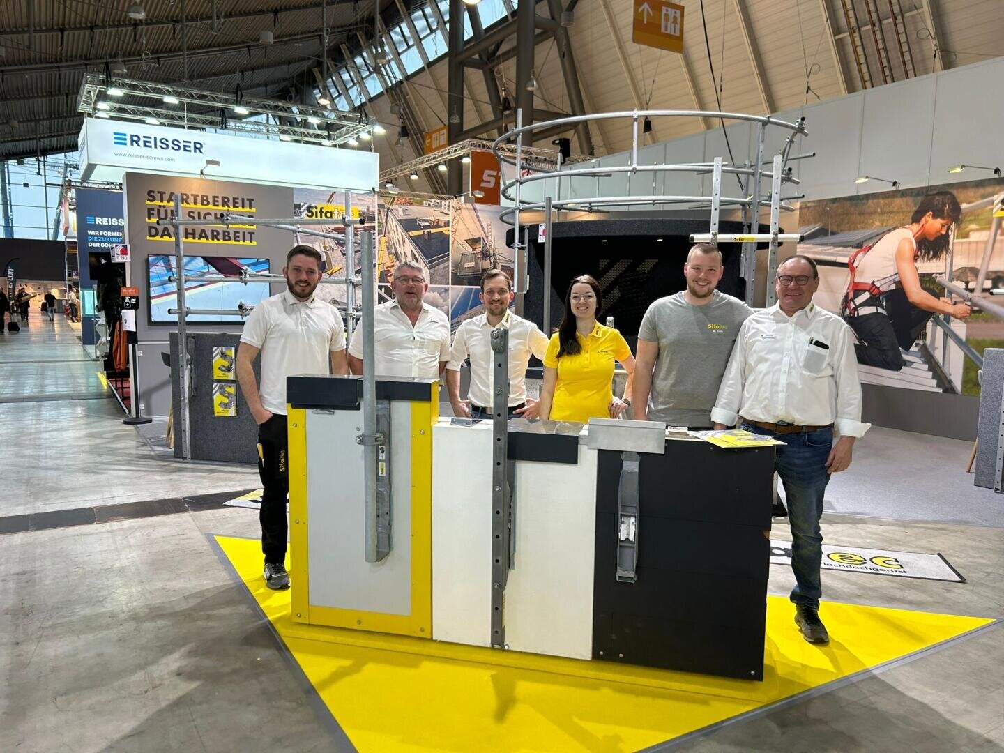 Fünf Personen stehen lächelnd hinter einem gelb-schwarzen Ausstellungsstand auf einer Indoor-Messe, im Hintergrund sind Baumaschinen, Banner und eine hohe Decke zu sehen.