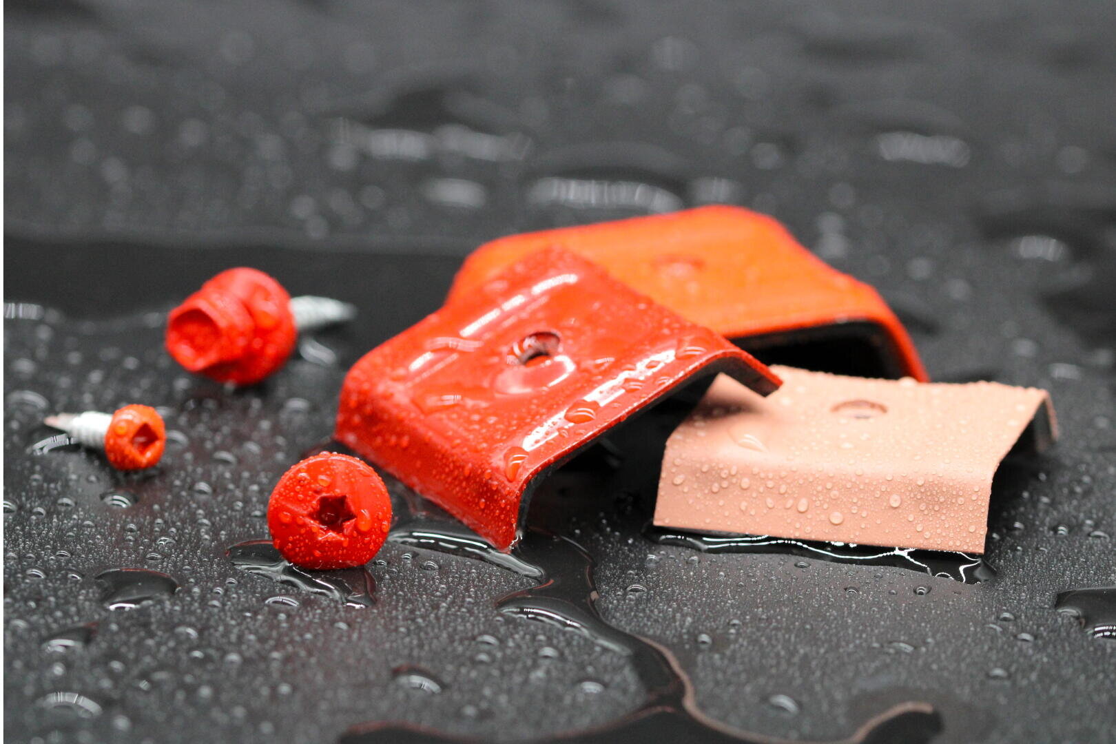 Mehrere orangefarbene und beigefarbene Kunststoffkappen, teilweise mit Schrauben, liegen auf einer nassen schwarzen Oberfläche, die mit Wassertropfen bedeckt ist. Die Objekte und die Tröpfchen erzeugen ein reflektierendes, glänzendes Aussehen.