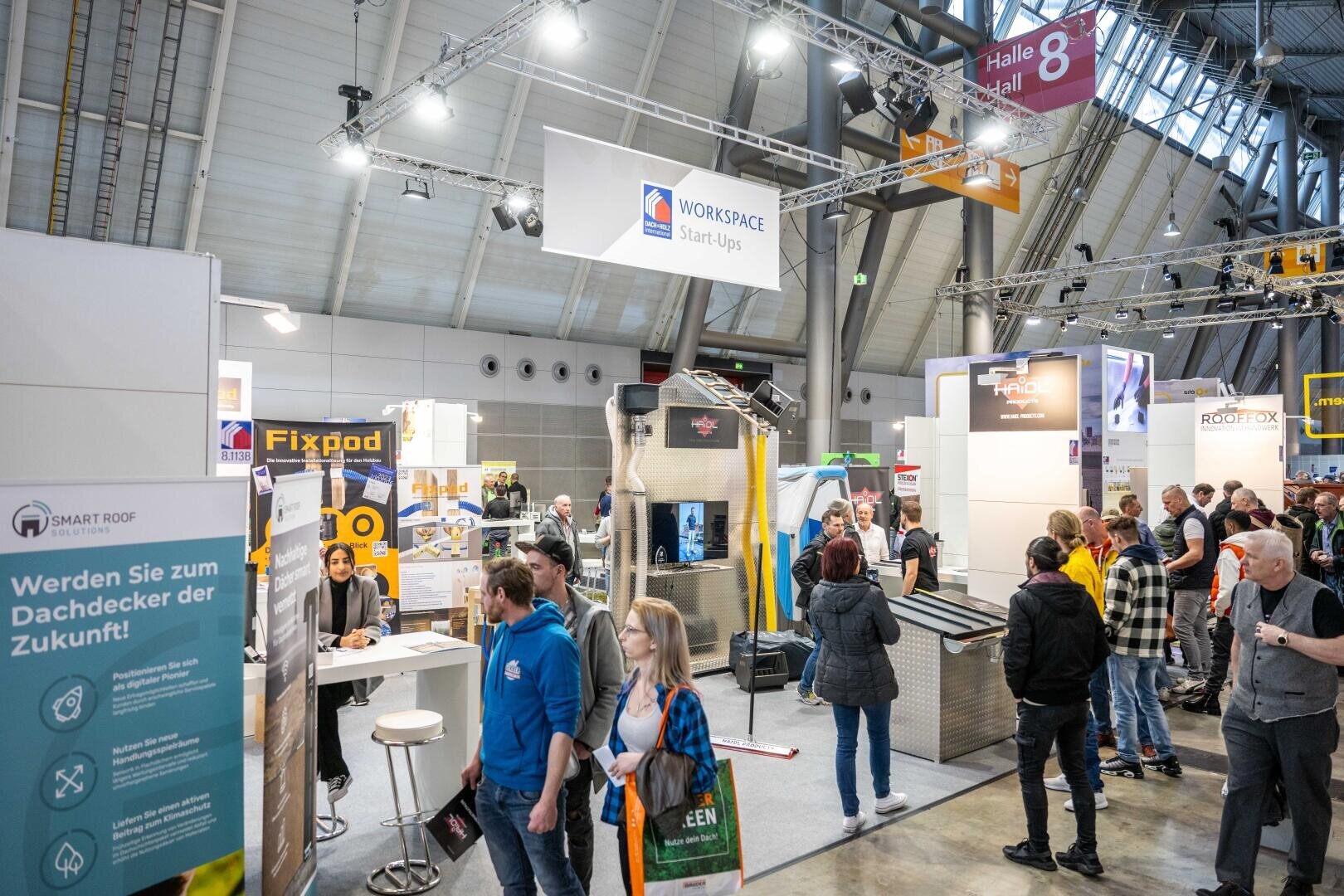 People visit a busy indoor trade fair with various stands and technology exhibitions. Signs for Workspace, Smart Roof and Fixpod are on display and visitors are walking around, chatting and looking at the products.