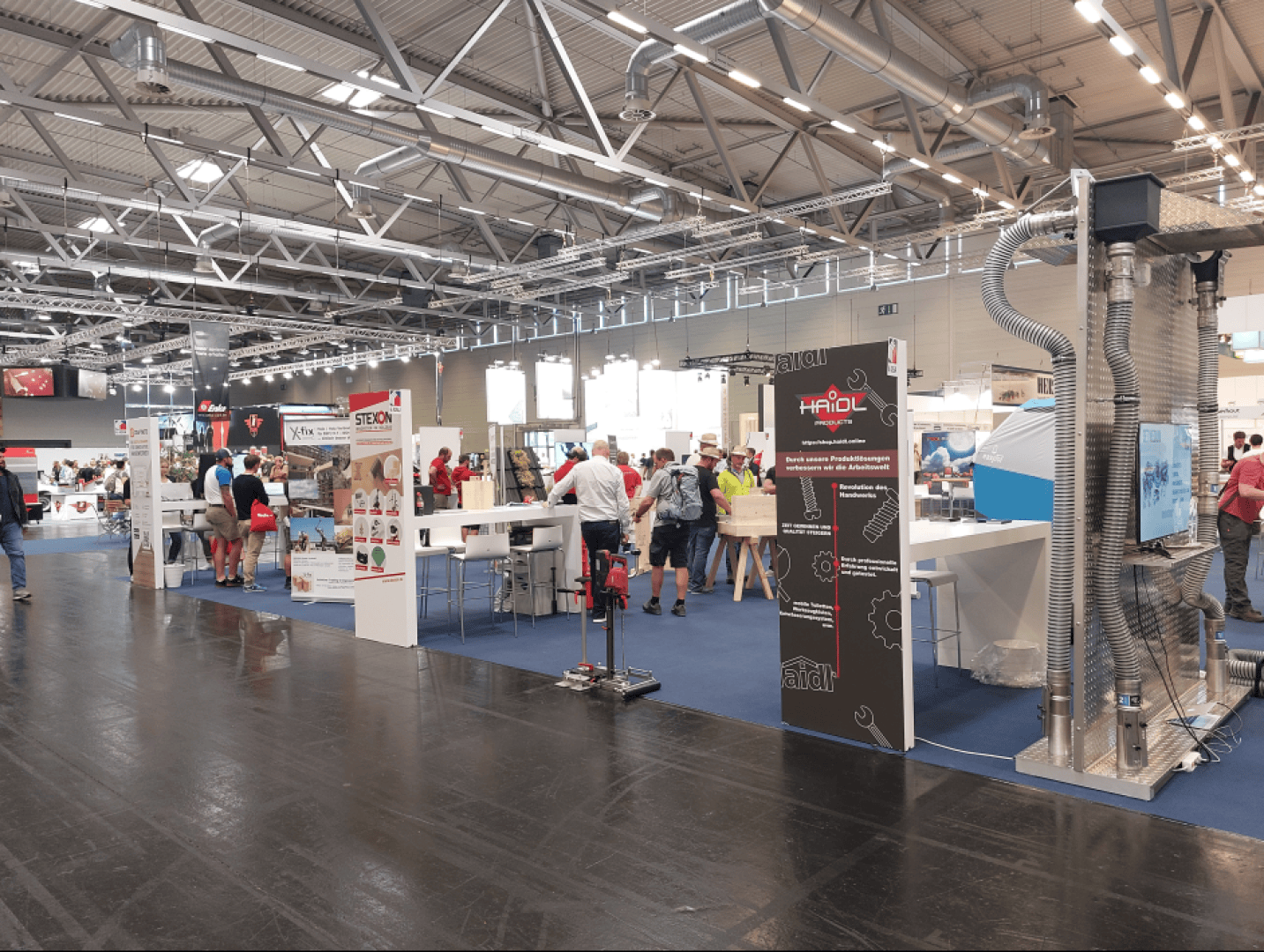 Eine Ausstellungshalle mit mehreren Ständen, Display-Ständen und Menschen, die gehen oder sich unterhalten. Der Bereich hat hohe Decken, industrielle Beleuchtung und Banner mit Firmenlogos und Produktinformationen.