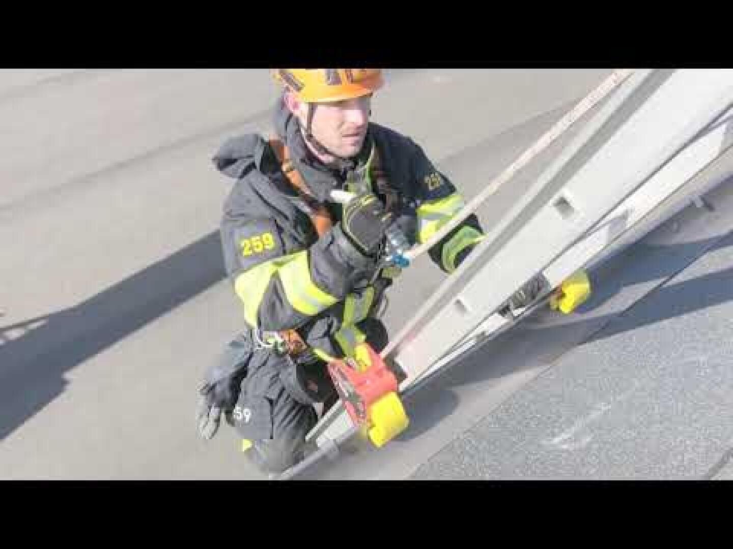 A firefighter in full gear and helmet climbs a ladder onto a rooftop during daylight. He is wearing a harness and carrying equipment, with the number 259 visible on his sleeve.