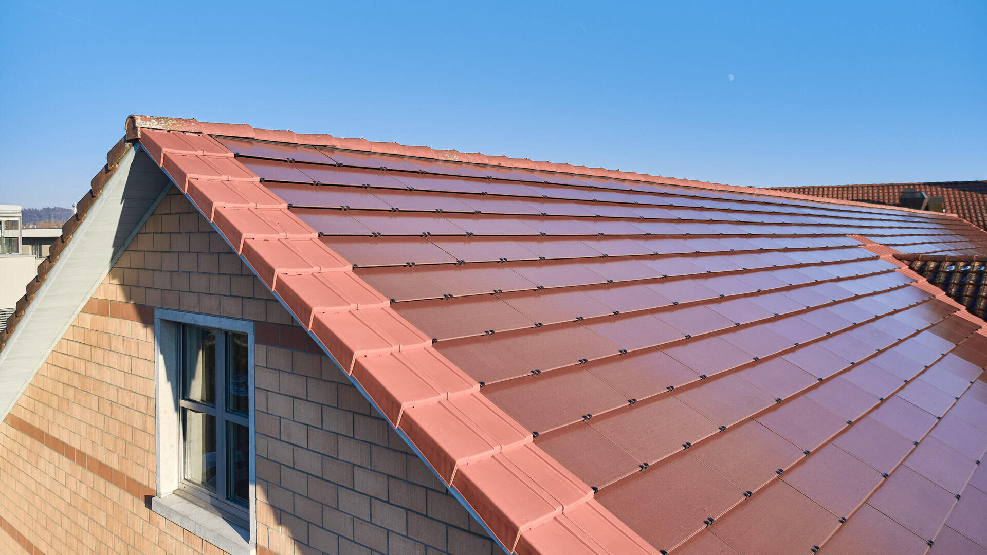 A pitched roof covered with red solar tiles can be seen under a clear blue sky. The outer wall is clad with beige and brown tiles, and a small window can be seen on the left-hand side.