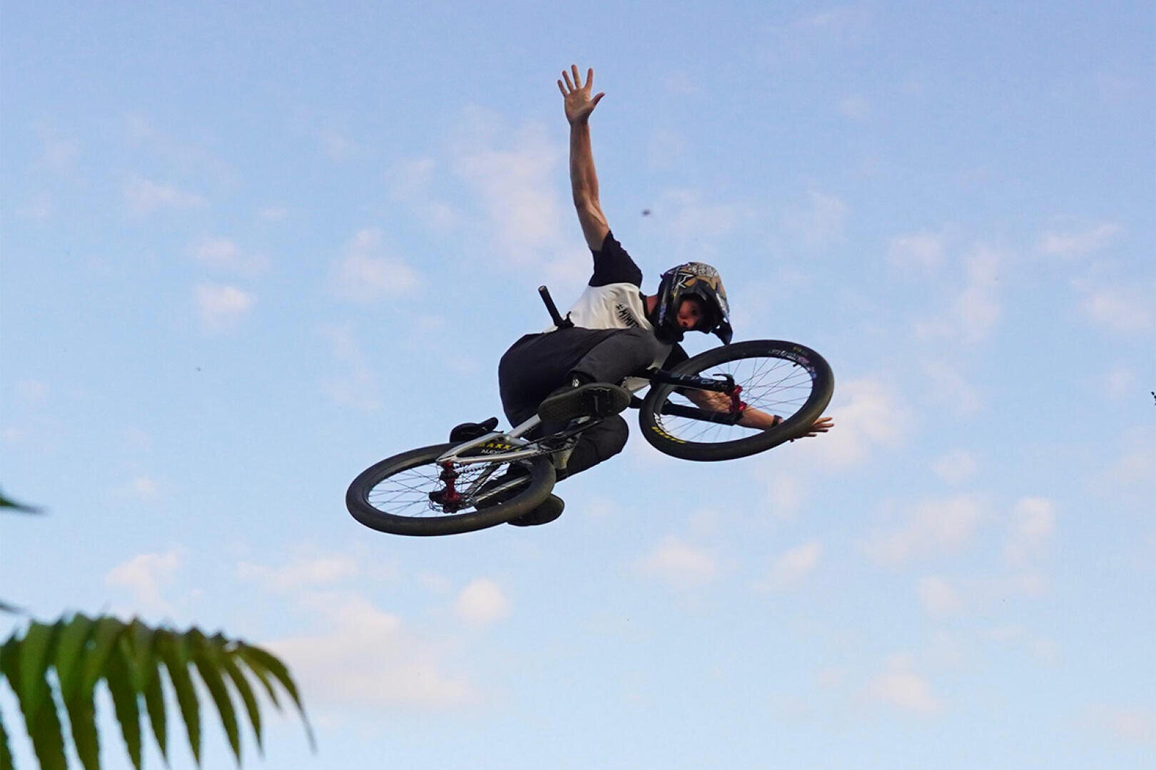 A person wearing a helmet performs a trick on a BMX bike, in the air with an outstretched arm in front of a blue sky with scattered clouds. Green leaves can be seen in the corner of the picture.