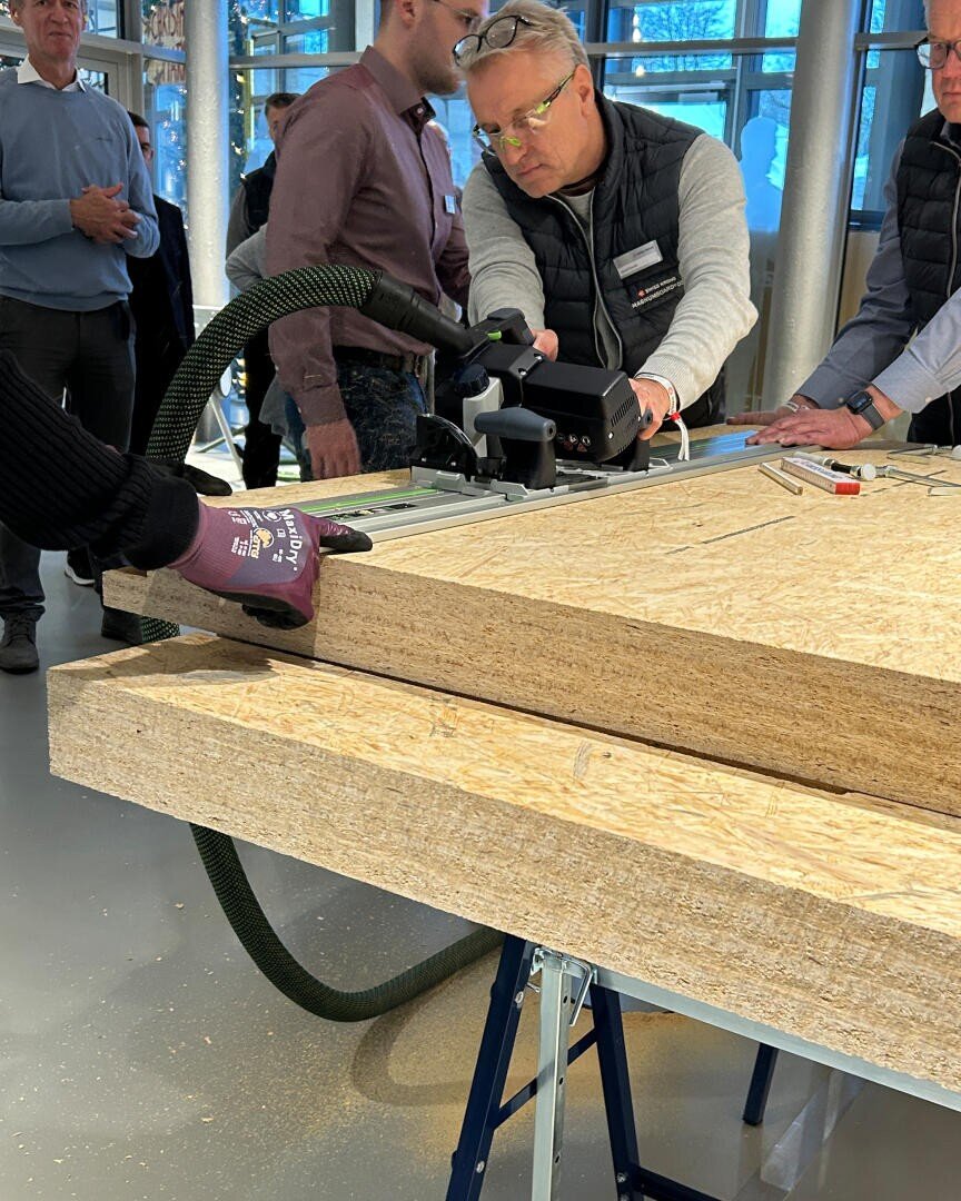 Eine Gruppe von Personen sieht zu, wie ein Mann mit einer Motorsäge ein dickes Holzbrett auf einer Werkbank zuschneidet. Der Arbeitsbereich befindet sich in einem Innenraum, und es werden Schutzhandschuhe und Schutzbrillen getragen.