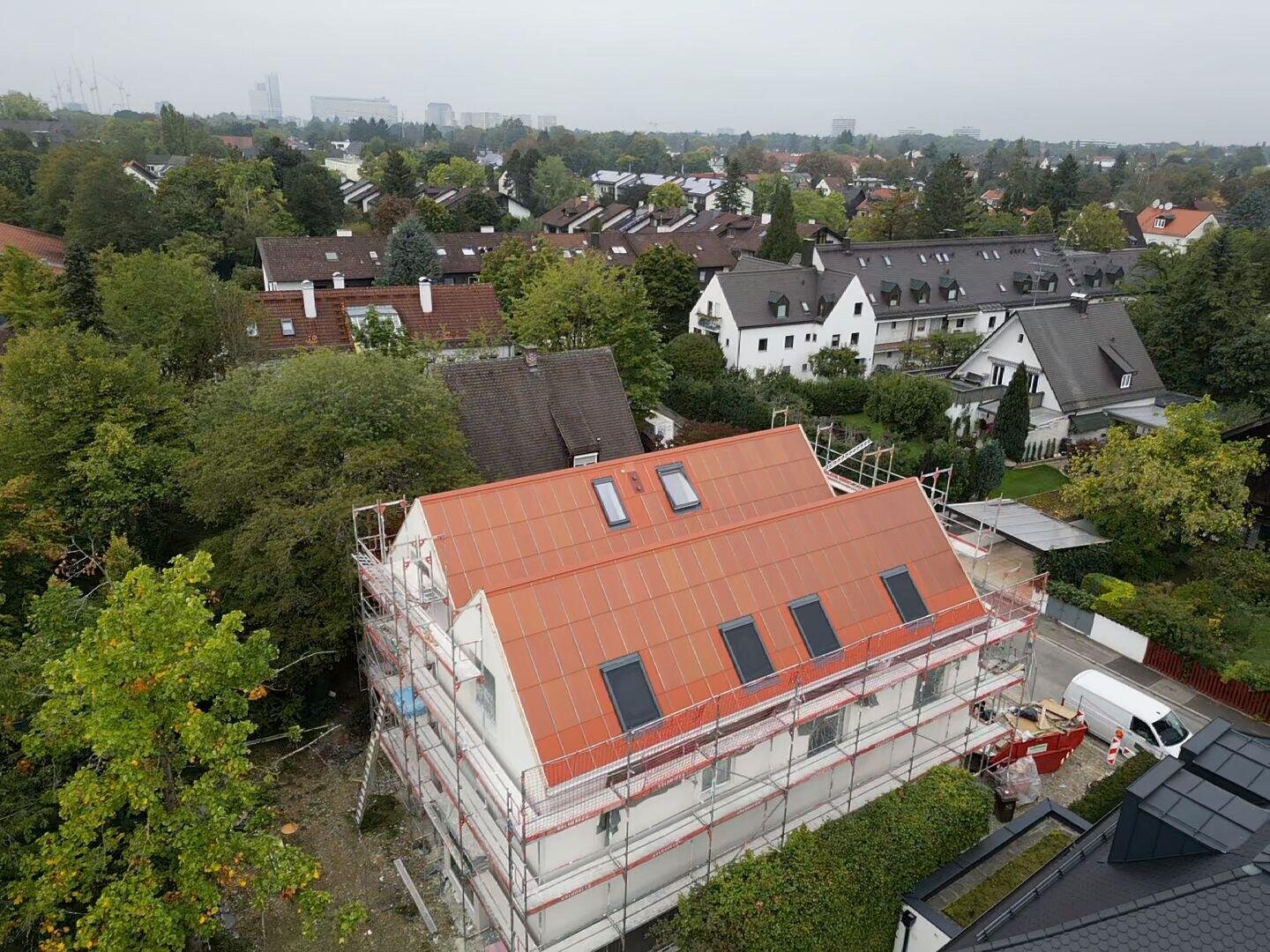 Luftaufnahme eines im Bau befindlichen Hauses mit Baugerüst und einem teilweise fertiggestellten roten Dach, umgeben von Bäumen und Nachbarhäusern in einem Vorort.