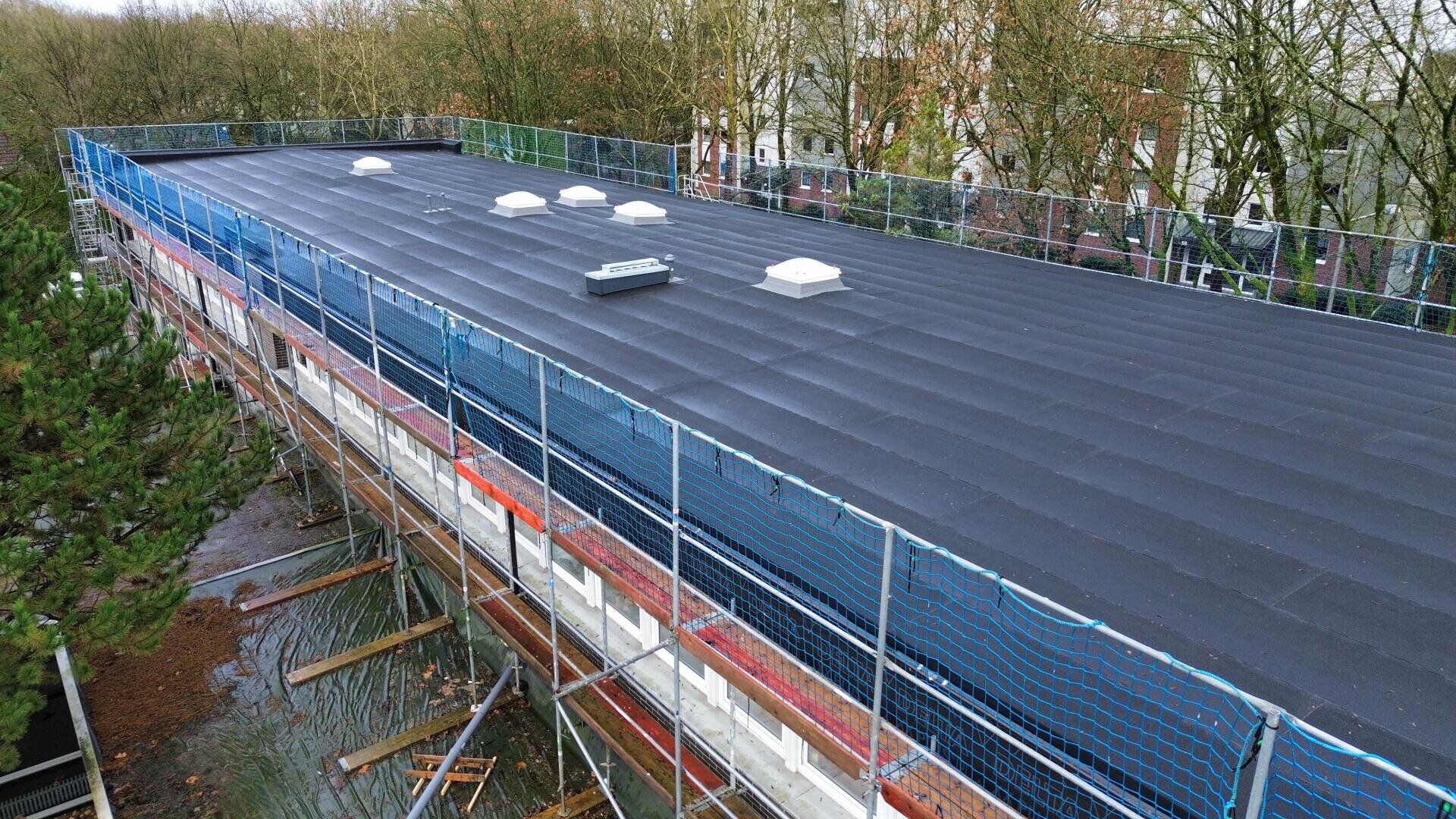 A large flat roof covered with black waterproof material, surrounded by blue safety nets and scaffolding, with several white skylights; trees and buildings can be seen in the background.