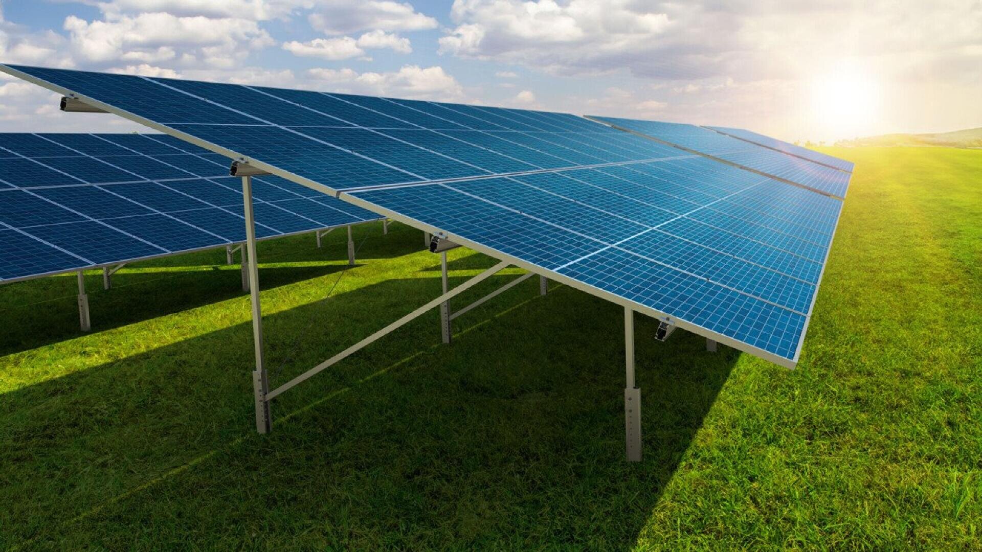 Rows of blue solar panels installed on metal frames stand in a green grassy field under a partly cloudy sky, with the sun shining brightly in the background.
