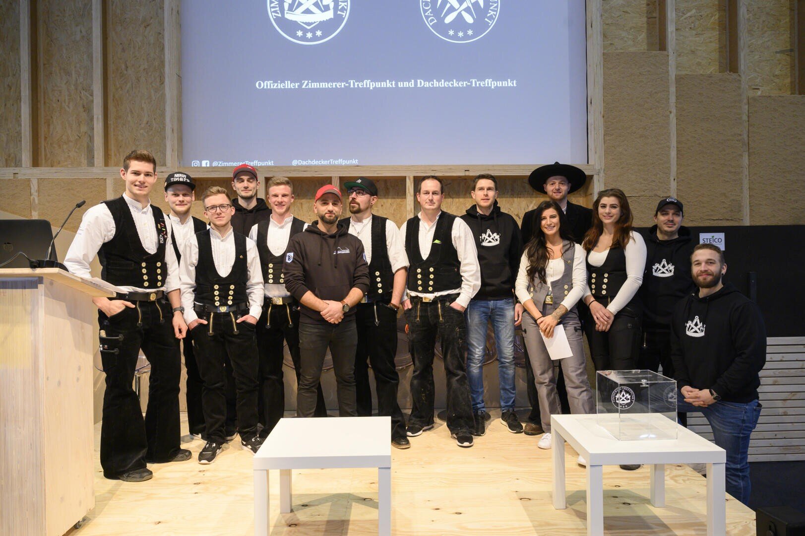 Eine Gruppe von Menschen, hauptsächlich Männer in traditioneller Tischlerkleidung, posiert auf einer Holzbühne für ein Foto. Dahinter ist ein Bildschirm mit Logos und deutschem Text zu sehen. Zwei Frauen und einige leger gekleidete Männer sind ebenfalls in der Gruppe.