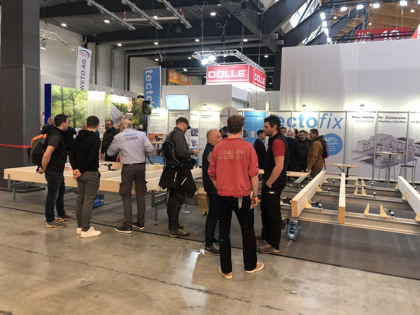 Eine Gruppe von Menschen steht um eine Holzbearbeitungsausstellung herum und unterhält sich, während im Hintergrund Holzbalken, Informationsposter und Banner zu sehen sind.