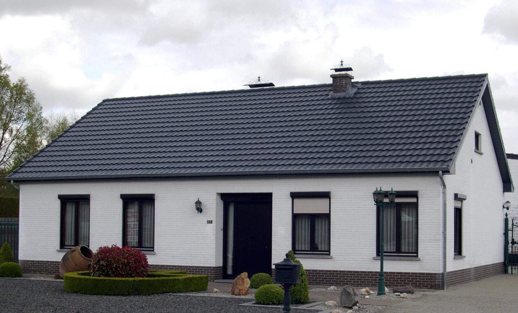 Ein einstöckiges weißes Haus mit einem dunkelgrauen Ziegeldach, schwarz umrandeten Fenstern, einer zentralen schwarzen Tür und einem kleinen, ordentlich angelegten Vorgarten mit Sträuchern und einem dekorativen Tontopf.