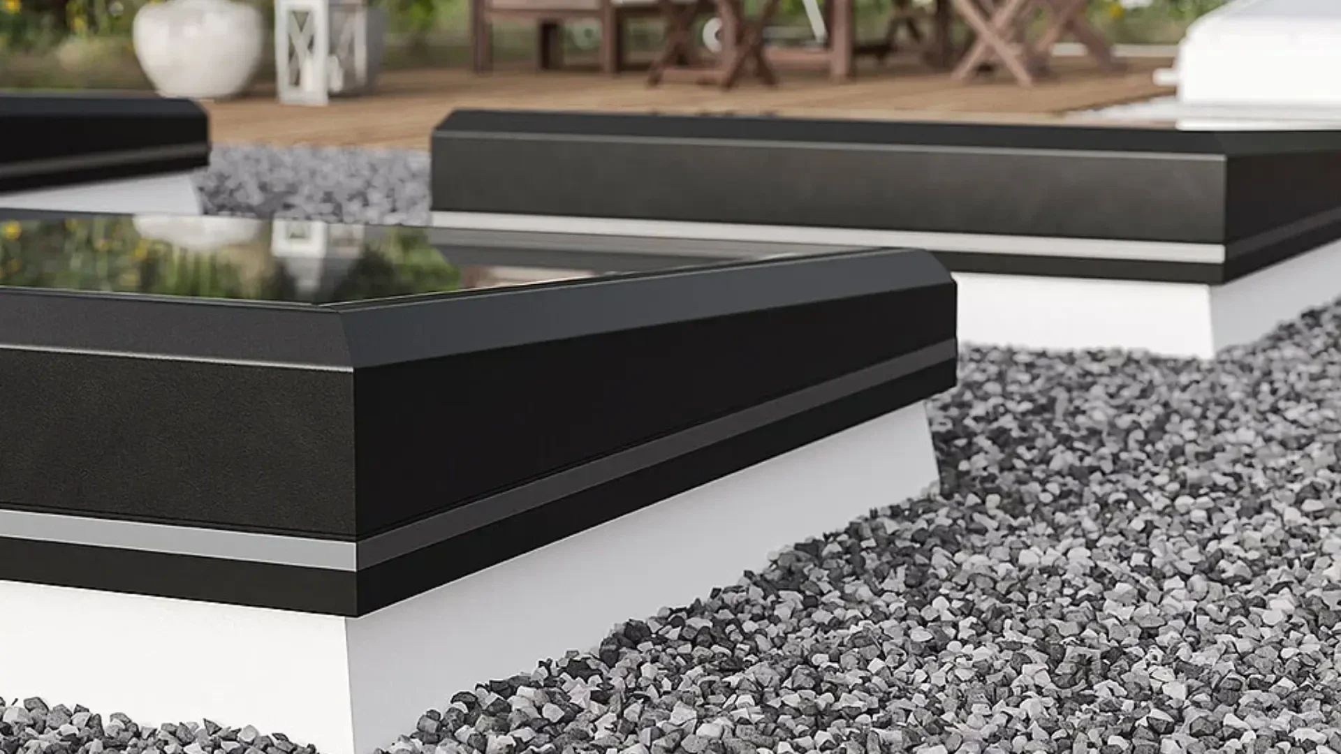 Nahaufnahme moderner schwarzer Oberlichter mit Glasplatten auf einem flachen, mit Kies bedeckten Dach. Im unscharfen Hintergrund sind eine Holzterrasse und Gartenmöbel im Freien zu sehen.