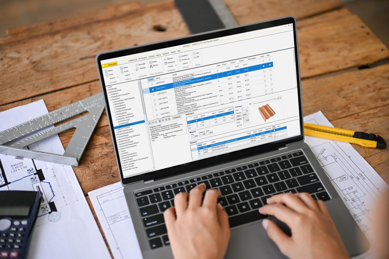 Eine Person arbeitet mit einem Laptop, auf dem eine Software für die Verwaltung von Bauprojekten angezeigt wird, an einem hölzernen Schreibtisch mit Bauplänen, einem gelben Universalmesser, einem Metalllineal und einem Taschenrechner in der Nähe.