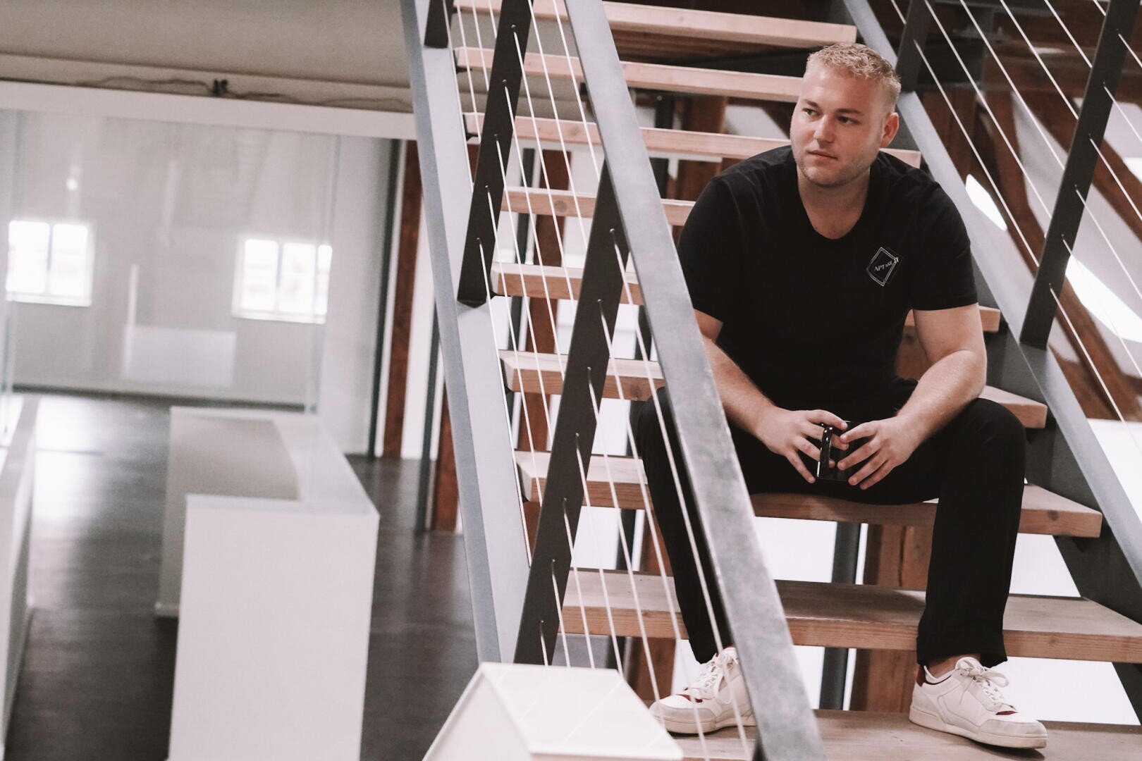 Ein Mann in schwarzem T-Shirt und weißen Turnschuhen sitzt auf einer modernen Holztreppe in einem Innenraum, hält einen Gegenstand in den Händen und blickt nachdenklich zur Seite.