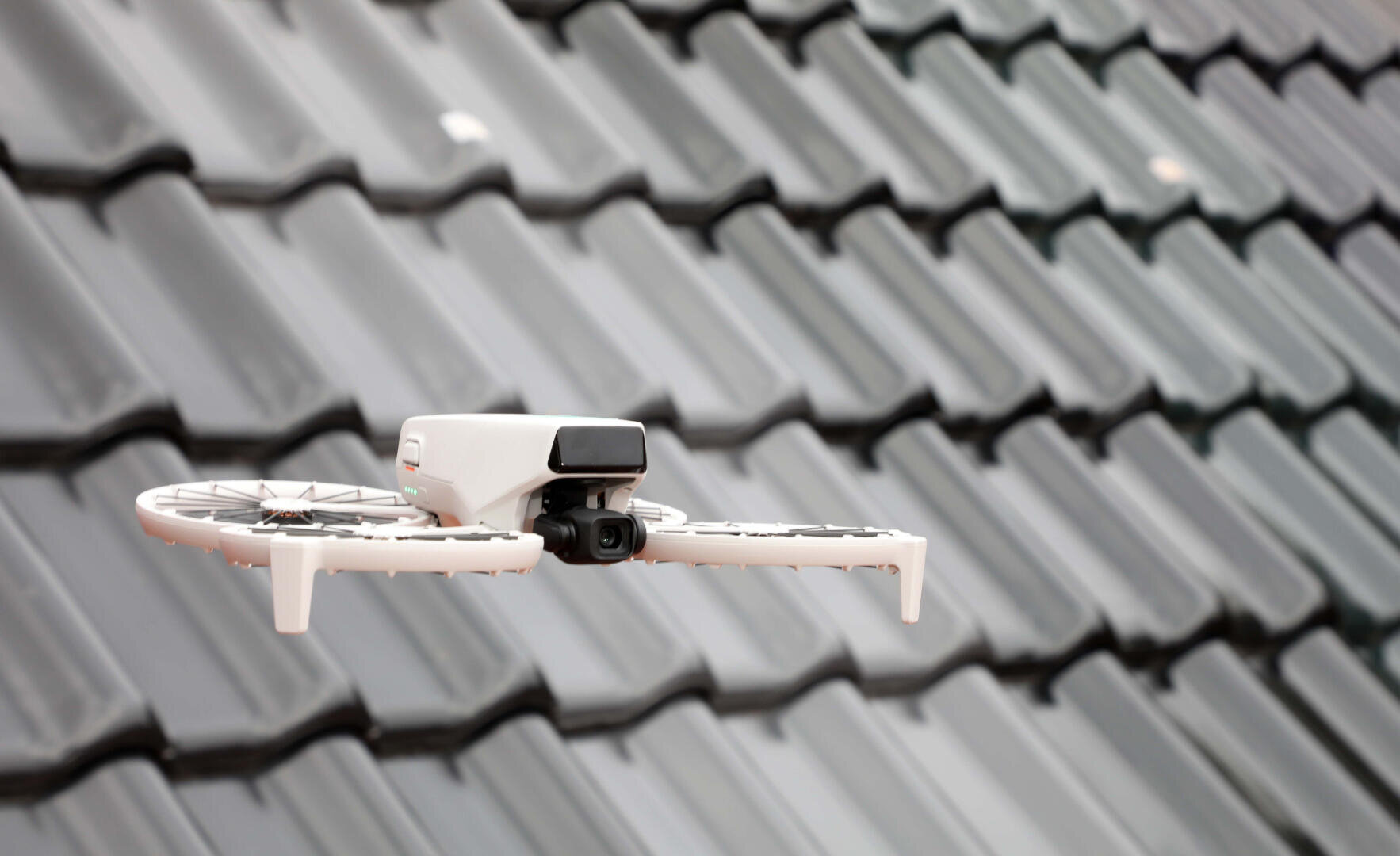 Eine kleine weiße Drohne mit einer Kamera fliegt vor einem grauen Ziegeldach, aufgenommen in der Luft bei Tageslicht.