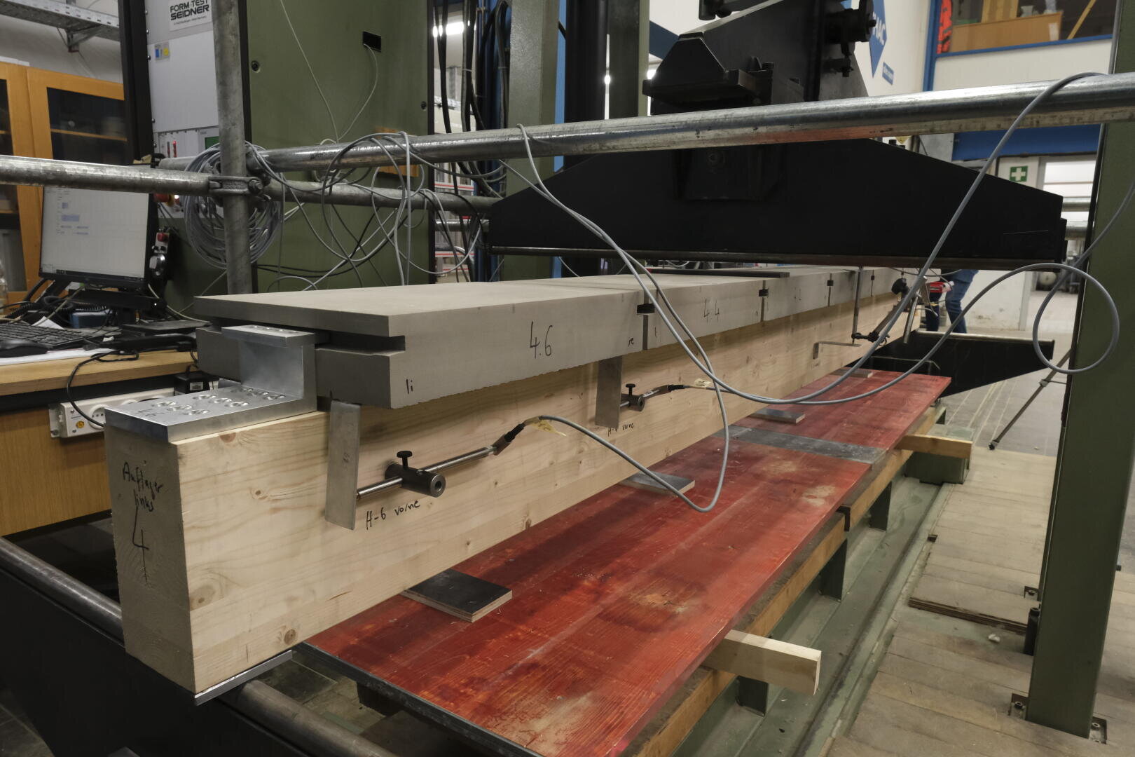 Ein Laboraufbau zeigt einen großen, mit Metallplatten verstärkten und mit mehreren Sensoren und Kabeln ausgestatteten Holzbalken, der auf einer roten Werkbank für Strukturprüfungen positioniert ist.