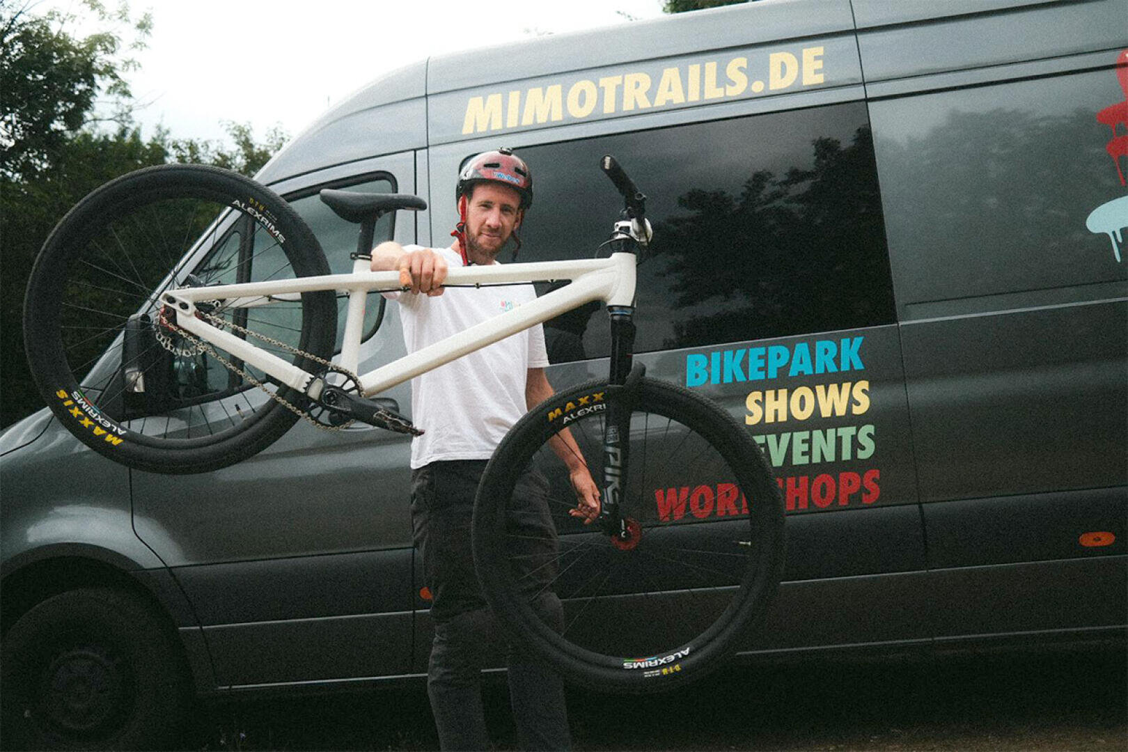A man in a helmet holds up a white mountain bike by the frame and stands in front of a dark van with the words MIMOTRAILS.DE and colorful words like BIKEPARK, SHOWS, EVENTS and WORKSHOPS on the side.