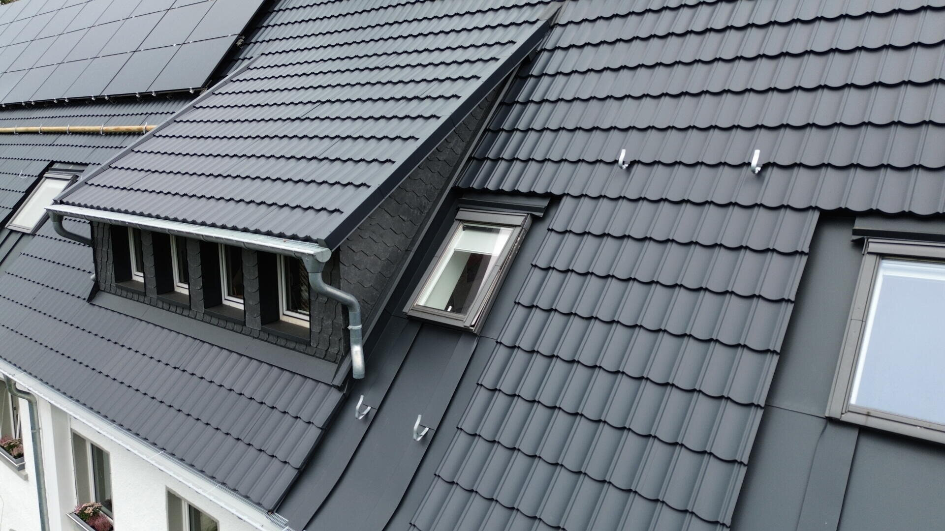 Nahaufnahme eines modernen Daches mit dunkelgrauen, überlappenden Metalldachziegeln, Sonnenkollektoren und Dachfenstern. Das Dach verfügt über eine Dachgaube und ein Dachrinnensystem.