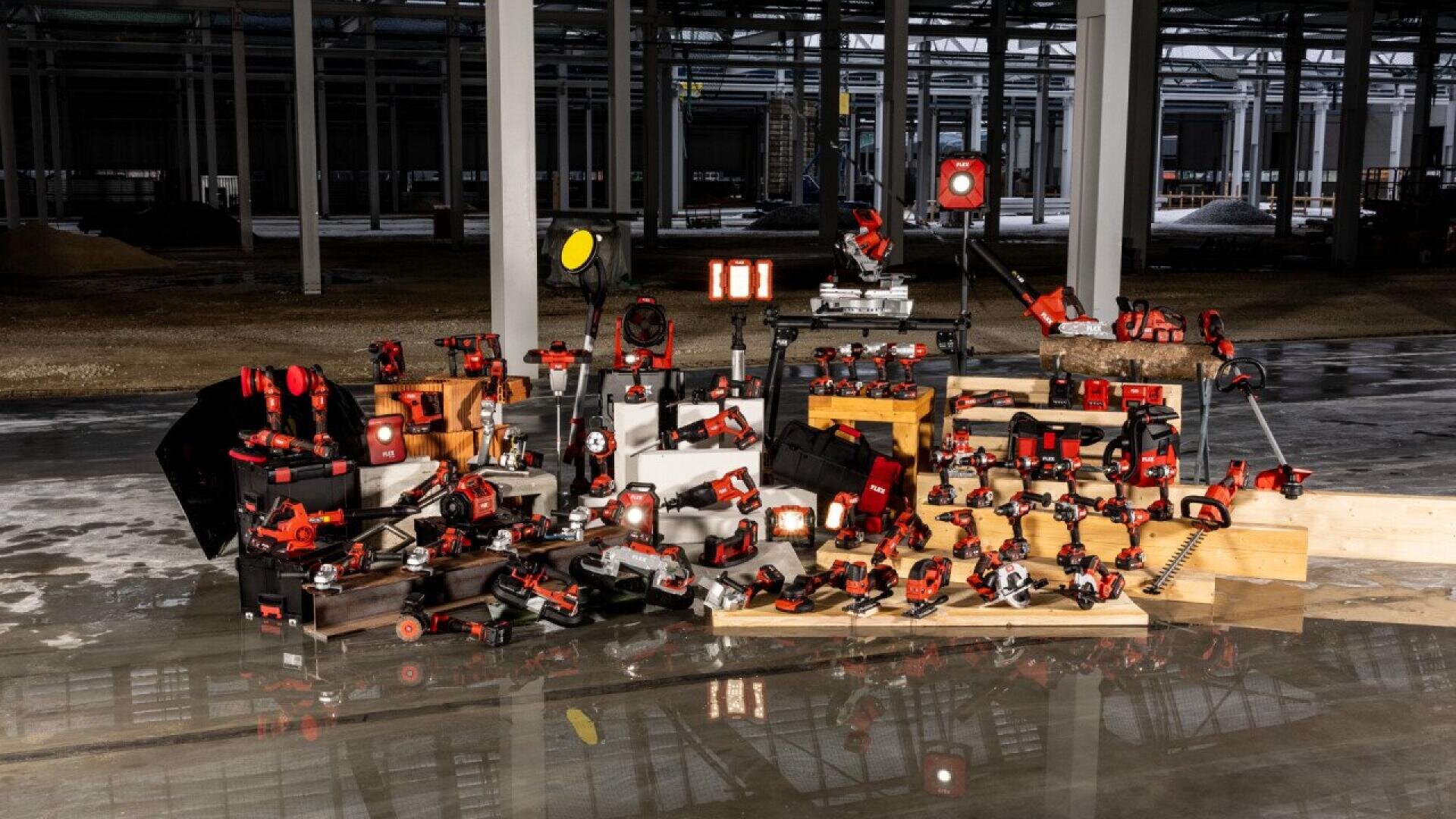 A large assortment of red and black power tools and equipment is displayed on wooden crates and stands inside an unfinished industrial building with concrete floors and metal beams.