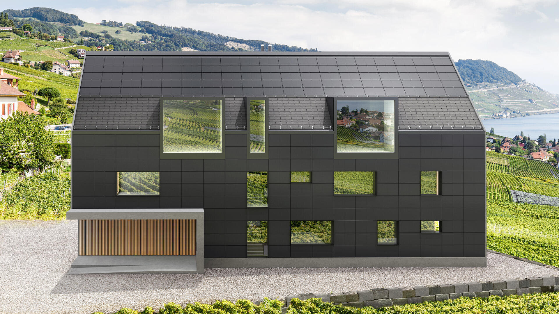 Ein modernes Haus mit schwarzen quadratischen Paneelen und großen Fenstern steht in einer Weinbergslandschaft, in der im Hintergrund unter einem bewölkten Himmel Hügel und ein Gewässer zu sehen sind.