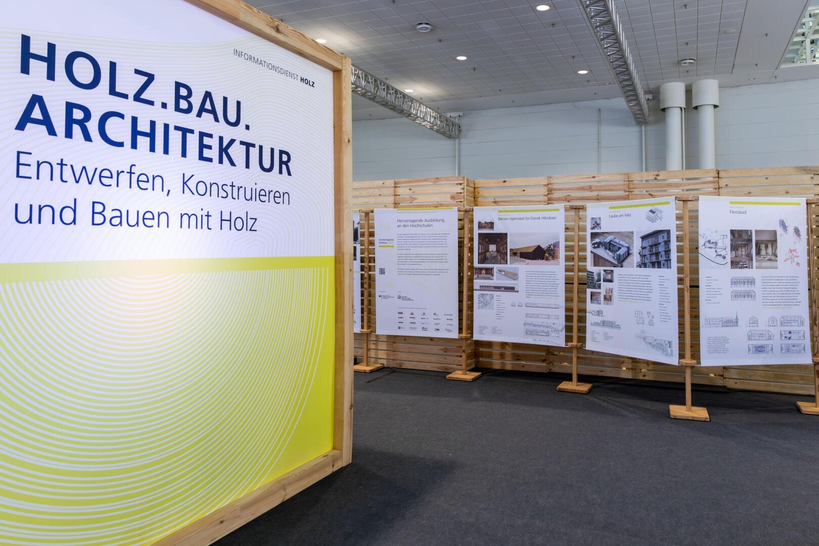 Ausstellungstafeln zeigen Informationen und Bilder zum Thema Holzbau und Architektur. Auf dem Hauptschild steht HOLZ.BAU. ARCHITEKTUR - Entwerfen, Konstruieren und Bauen mit Holz auf Deutsch.