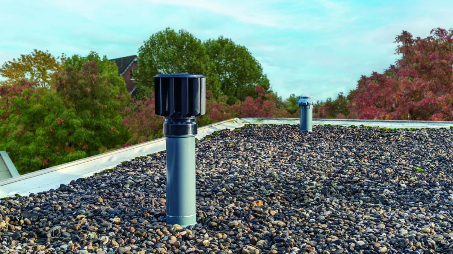 Ein mit Kies bedecktes Flachdach mit zwei Entlüftungsrohren, eines davon mit einer schwarzen zylindrischen Kappe, umgeben von grünen und roten Bäumen unter einem teilweise bewölkten Himmel.