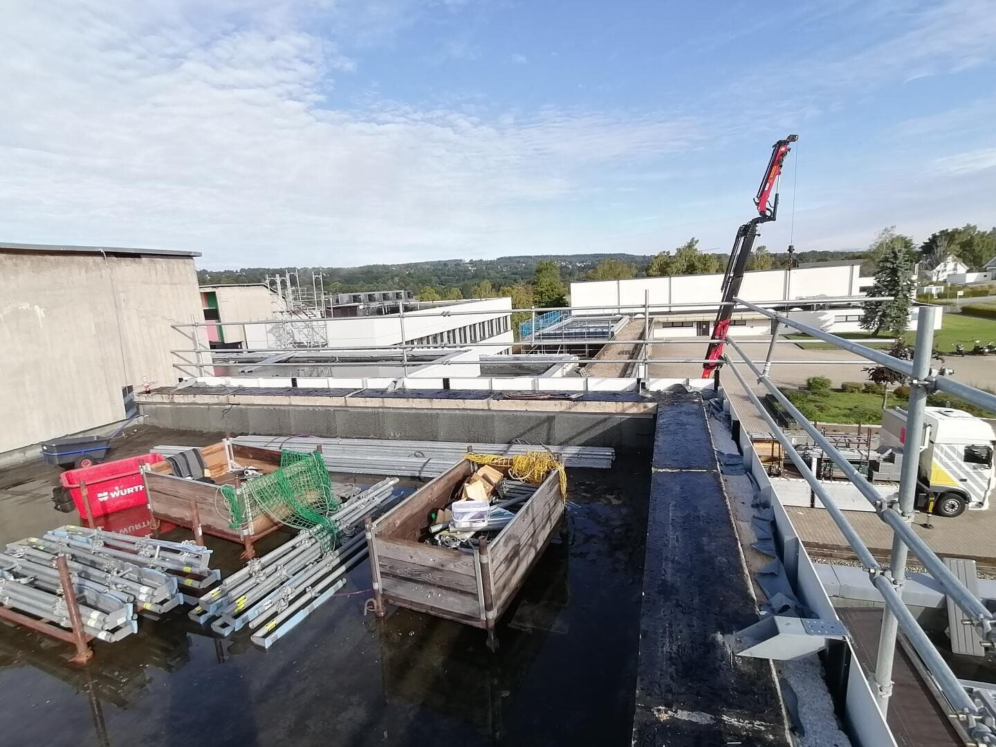 Baumaterialien und Schutt sind auf einem flachen Dach verstreut, das von Metallgeländern umgeben ist. Ein Kranarm und ein weißer Lastwagen sind zu sehen. Im Hintergrund sind Gebäude und eine grüne Landschaft unter einem teilweise bewölkten Himmel zu sehen.
