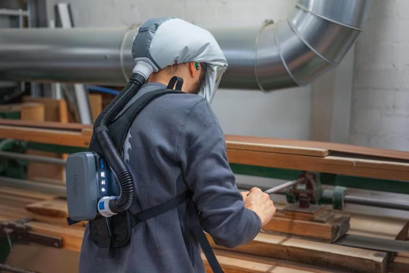 Eine Person mit Schutzausrüstung, einschließlich Haube und Atemschutzgerät, arbeitet mit Holzbrettern in einer Werkstatt, wobei im Hintergrund große Lüftungsrohre zu sehen sind.